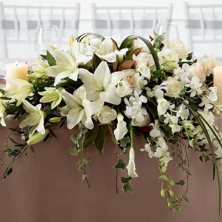 White Linen Arrangement - The White Linen Arrangement speaks to the true nature of wedding elegance. White Dendrobium Orchids, Asiatic Lilies, mini calla lilies, and roses are accented with an assortment of lush greens and arranged to add to the decor of the wedding party's table with its sweet sophistication.  Approx. 22"H x 40"W.