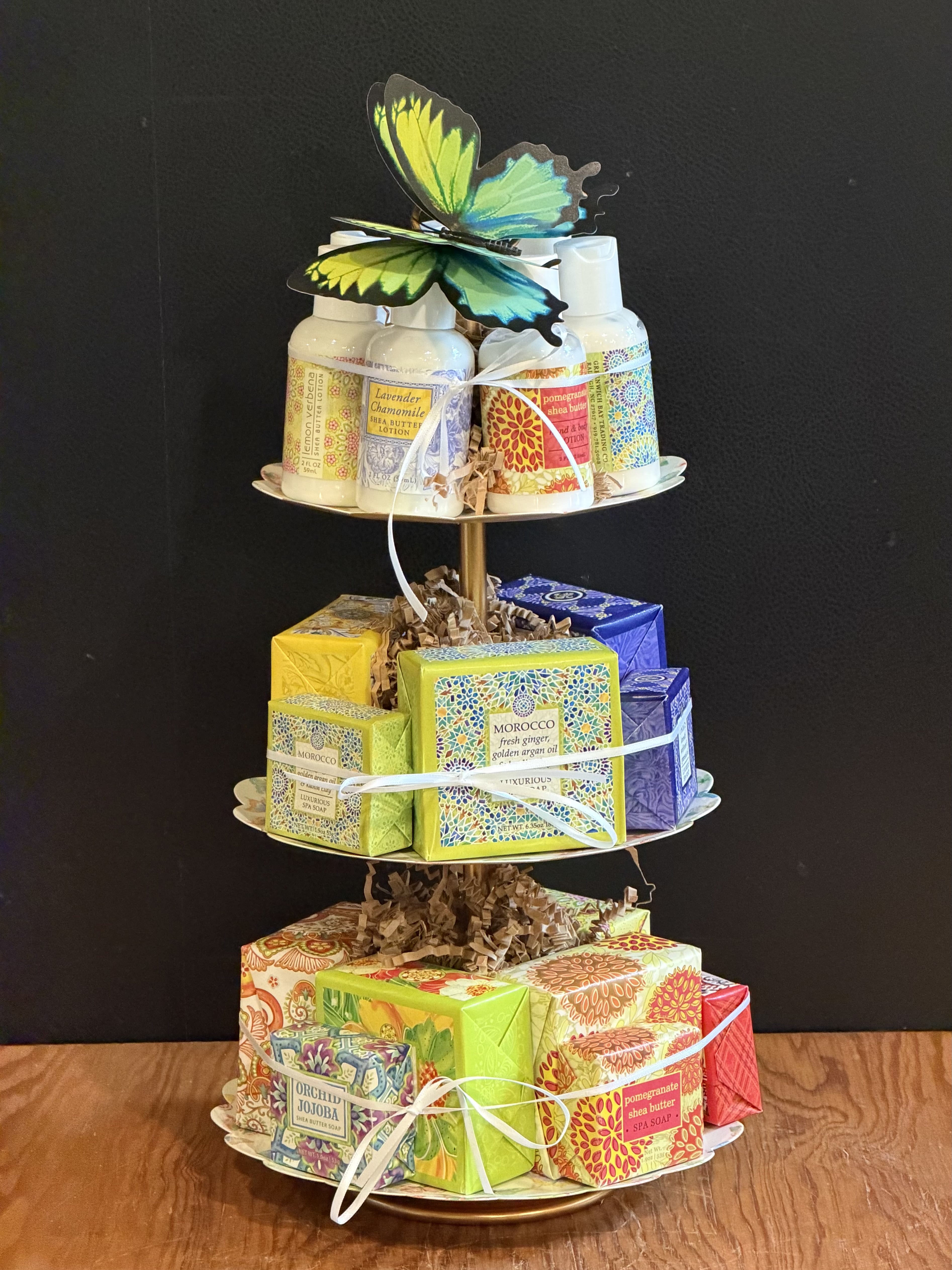Tower Of Spa Essentials - A bright, cheerful spa-style gift tower that reads like a thoughtful pampering present - perfect as a thank-you gift, a birthday surprise, or a "thinking of you" treat for someone who loves little luxuries. This three-tiered spa arrangement pairs boxed soaps and lotion bottles in mixed colors (yellow, purple, red, green and blue) arranged on scalloped white plates, topped with a playful butterfly decoration to make the recipient feel truly spoiled.  The display sits on a tiered metal stand with a slim gold post and ring handle, featuring three white decorative plates printed with tiny multicolored butterflies. Each tier is finished with shredded paper filler, neatly tied with white ribbon, and includes soap boxes and lotion bottles grouped for visual balance - a lively mix of yellow, purple, white and pink tones that reads joyful and celebratory. The gold handle and decorative butterfly topper give it a polished, gift-ready look.  Perfect for hostess gifts, bridal or baby shower favors, or a cheerful get-well or thank-you present, this tower is ready to set out as a centerpiece or place on a vanity for everyday indulgence. No special care is required beyond keeping the products dry and in a cool spot; order now for same-day pickup or delivery to make someone feel pampered the moment it arrives.
