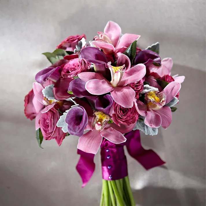 Delicacy Bouquet - Chic, stylish, and utterly romantic, this bridal bouquet is simply enchanting at every turn! Purple garden roses flaunt their many ruffled petals arranged amongst red mini calla lilies, plum min calla lilies, and pink cymbidium orchids, perfectly accented with Dusty Miller stems for an alluring affect.Tied at the stems with a bold purple satin ribbon to add even further appeal and sophistication to this already stunning design, this bouquet is ready to make it's entrance on your wedding day.  Bouquet is approximately 13" H x 13" W