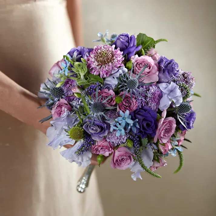 Lavender Garden Bouquet - Enchanting and romantic, this stunning wedding bouquet brings together the look of your ladies with a certain panache you want to convey on your special day. Lavender sweet pea, purple Veronica, lavender scabiosa, lavender spray roses, purple double lisianthus, sea holly, tweedia, and ornamental kale are arranged to perfection to create a stunning display. Affixed with a silver metallic bouquet holder to add to the appeal and sophistication of your wedding party's look, this bouquet has a classic elegance that will make your guests take notice.  Bouquet is approximately 10" H x 8" W
