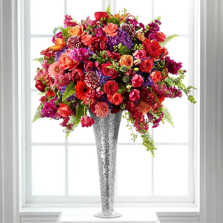 Flower Jeweled Arrangement - Simply dazzling and rich in color and design, this gorgeous arrangement will transform your church, outdoor venue, or reception hall into an unforgettable experience for you and your wedding guests. Salmon roses, green trick dianthus, hot pink tulips, burgundy peonies, purple hydrangea, hot pink roses, red mokara orchids, pink gilly flower, magenta gilly flower, pink mini carnations, coral roses, orange gerbera daisies, burgundy and white dahlias, red garden roses, coral dahlias, lavender mini calla lilies, burgundy Asiatic Lilies, globe thistle, orange Asiatic Lilies, burgundy mini calla lilies, Boston Fern fronds, and bittersweet branches are arranged perfectly within a tall sparkling vase to add glamour and color to wherever you choose to place it. A grand way to celebrate your love!  Approx. 43"H x 32"W.