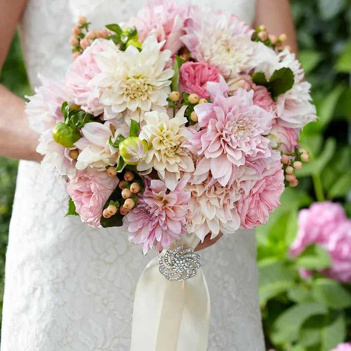 Heart's Promise Bouquet - Frilled, feminine, and perfectly elegant, this bridal bouquet is an absolute dream. Pink garden roses, known for their abundance of ruffled petals, are brought together with blush and cream dahlias, and peach hypericum berries to create an exquisite arrangement. With cream satin ribbon tied at the stems brought together with a spiral brooch to create even more drama, this bridal bouquet is a sophisticated choice for the modern bride.  Bouquet is approximately 13" H x 11" W
