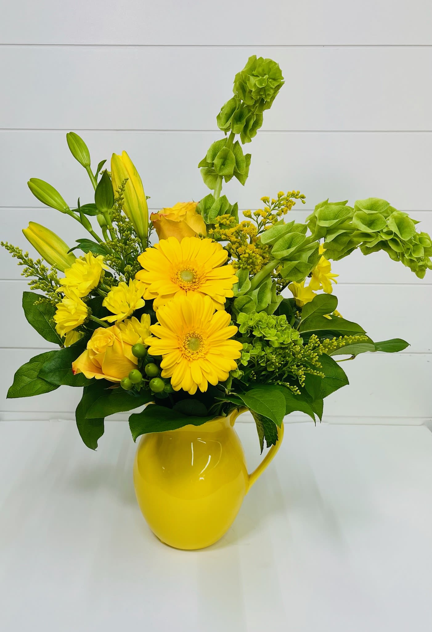 Mama Bella - Fresh, cheerful, and full of life, Mama Bella is a sunny celebration of soft beauty and natural charm. Designed in a bright yellow pitcher, this arrangement features a vibrant mix of yellow roses, classic white daisies, elegant lilies, and striking Bells of Ireland, all accented with lush greenery.  The combination of yellow and green tones creates a refreshing, uplifting feel—perfect for brightening someone’s home or sending a thoughtful reminder that they’re loved. With its airy, garden-inspired style and soft movement, Mama Bella brings warmth and light to any space.  The keepsake yellow pitcher adds a charming touch and can be repurposed long after the blooms have faded, making this gift both beautiful and lasting.  Perfect for Mother’s Day, birthdays, thank-you gifts, or simply because, Mama Bella is made for anyone who loves fresh color and effortless elegance.  Delivery Available In: Smyrna, Marietta, Mableton, Austell, and Atlanta