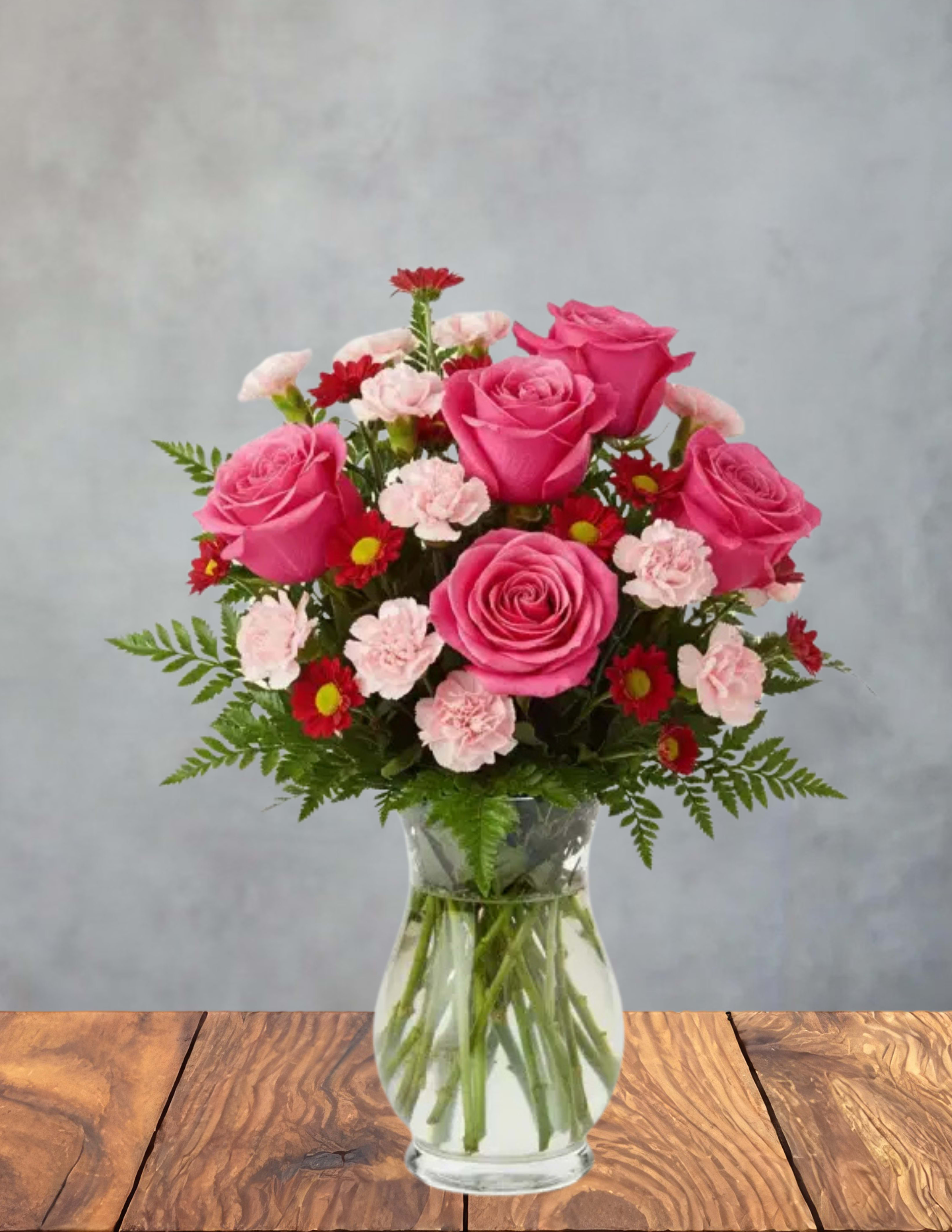Blossoming Heart - Bright pink roses take center stage in this cheerful, romantic flower arrangement-perfect as a birthday bouquet or an anniversary gift that reads as both heartfelt and joyful. The rounded, full shape and lively pink-and-red palette make it a lovely choice for love and romance, a "just because" surprise, or a tasteful thank you; its soft blush accents also allow it to offer gentle comfort for sympathy and funeral needs without feeling overwrought.  Clusters of bright pink roses are paired with pale pink carnations and small red daisy-like blooms, all tucked into airy fern foliage for a light, textured silhouette. The stems are visible through the clear glass vase, giving the piece a classic, fresh look that suits casual or formal settings. Size and proportions create a generous, full-top bouquet that makes the recipient feel truly celebrated.  Easy to place on a table, desk, or bedside, this arrangement arrives ready to brighten someone's day-whether marking a milestone, expressing gratitude, or saying you're thinking of them. For best longevity, change the water every couple of days and recut stems slightly when you refresh the vase.
