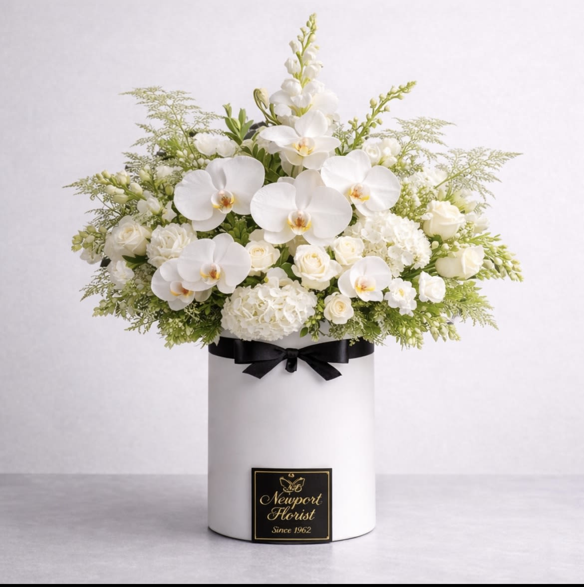 White Serenity By Newport Florist - A serene cluster of white phalaenopsis orchids, roses, and hydrangeas creates a timeless sympathy or wedding arrangement that will make the person you're thinking of feel calmly supported and truly seen. The white flower arrangement is presented in a tall white hat box-an elegant choice for a sympathy tribute or a refined bridal table accent-and finished with a contrasting black ribbon and a small brand plaque for a polished, formal look.  Soft, rounded hydrangea clusters and classic white roses provide lush volume while the long, arching Phalaenopsis orchids lend graceful height and a quietly luxurious focal point. Fine airy greenery fans the edges, giving the design a balanced silhouette and gentle texture against the crisp white palette with just a touch of black from the ribbon for visual contrast. The arrangement sits in a tall hat box, making it easy to place on a memorial table, altar, or reception surface.  Perfect as a sympathy expression, a wedding centerpiece, or a sophisticated gift to mark a solemn milestone, this arrangement reads as both elegant and comforting-an understated way to honor, celebrate, or console. Order with standard sizing in the tall white hat box; to preserve blooms, keep in a cool spot away from direct sunlight and mist the orchids lightly if air is dry.