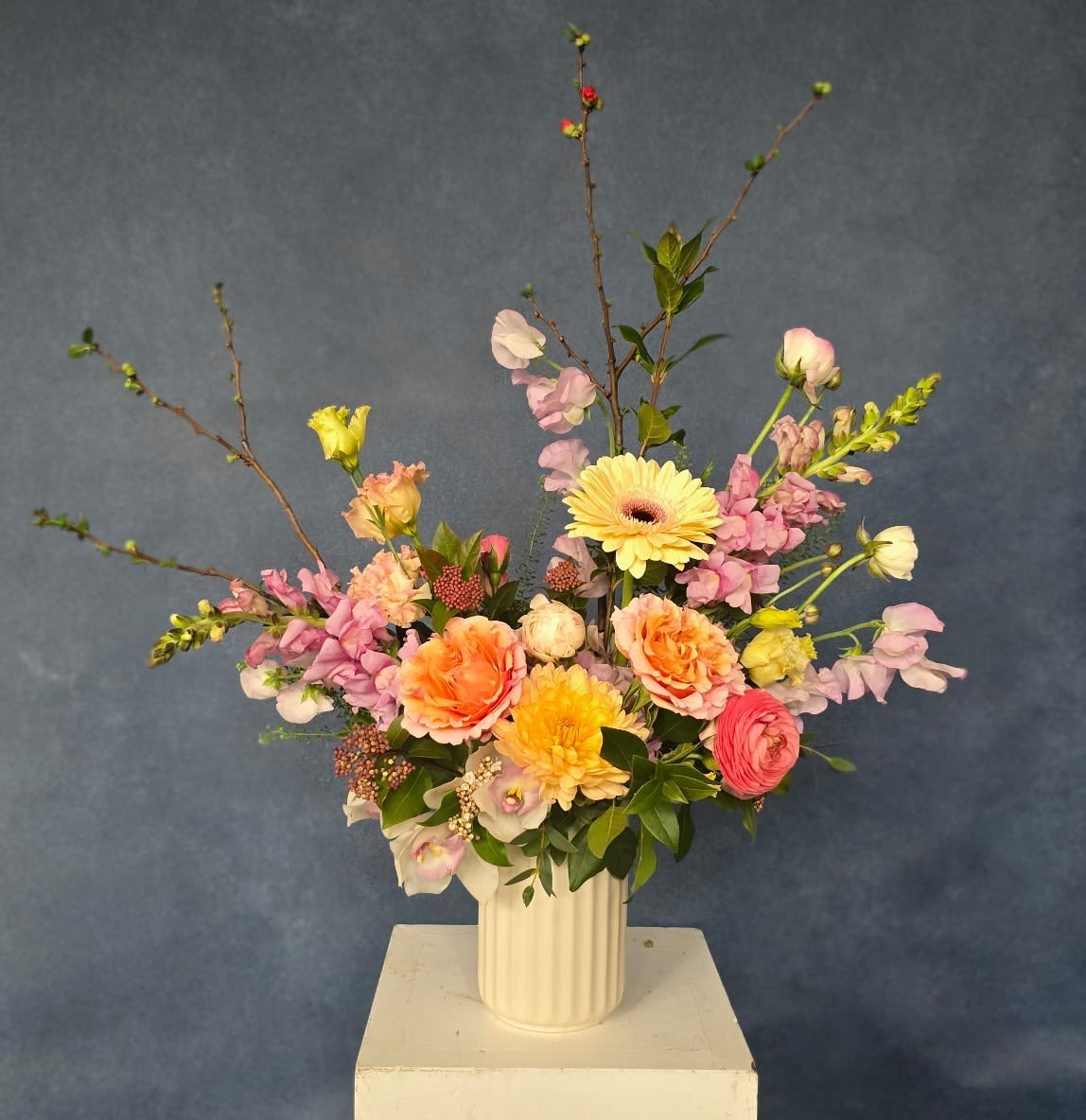 Seasonal Designers Choice - A soft, garden-style bouquet in a white ribbed ceramic vase, blooming with airy pastels that feel both romantic and effortlessly fresh. Perfect as a spring celebration bouquet, a cheerful everyday gift, or for a soft wedding-inspired moment, this seasonal designers choice features a loose, layered composition with branching stems that add height, movement, and a relaxed, handpicked look your recipient will notice.  Colors range through gentle pinks, peaches, buttery yellow, and ivory, arranged to create a delicate, expansive presence on a table or mantel. The ceramic vase gives it a clean, modern touch and makes the arrangement ready to display; include any color preferences in the special instructions and our team will tailor the selection to suit the occasion and the person you're celebrating.