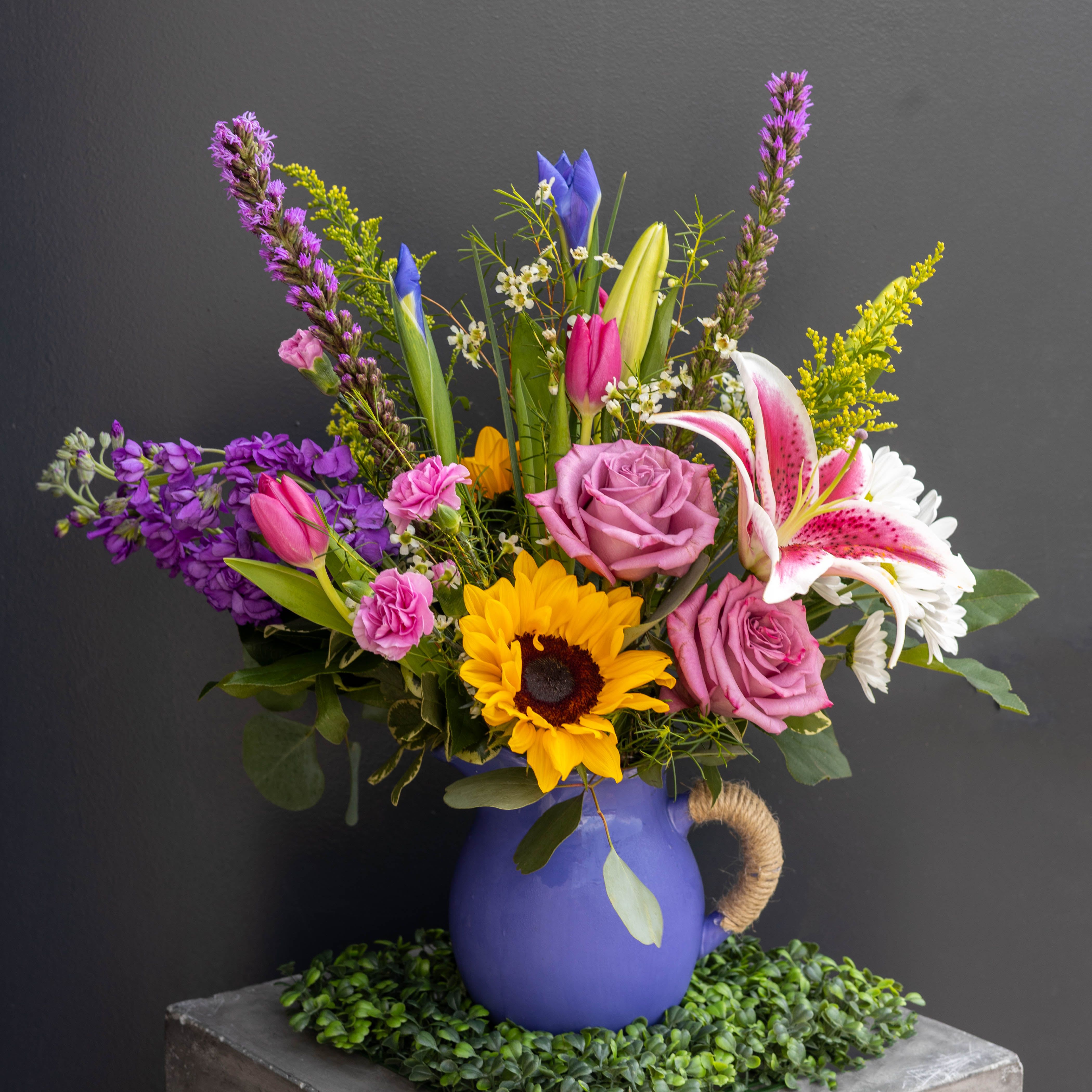 Berry Bliss - Bright, playful, and bursting with color, Berry Bliss is a joyful celebration of spring at its peak. This vibrant arrangement blends cheerful blooms in shades of sunny yellow, juicy pink, and fresh purple, creating a lively, fruit-inspired palette that instantly uplifts any space.  The arrangement is beautifully presented in a glossy ceramic pitcher-style vase in a rich periwinkle hue, complete with a softly curved silhouette and a rustic rope-wrapped handle for a touch of farmhouse charm. The unique design adds both character and warmth, making it a keepsake piece long after the blooms have faded.  Perfect for birthdays, celebrations, or just because, Berry Bliss is designed to make people smile. It’s bright, energetic, and full of personality...just like spring should be!