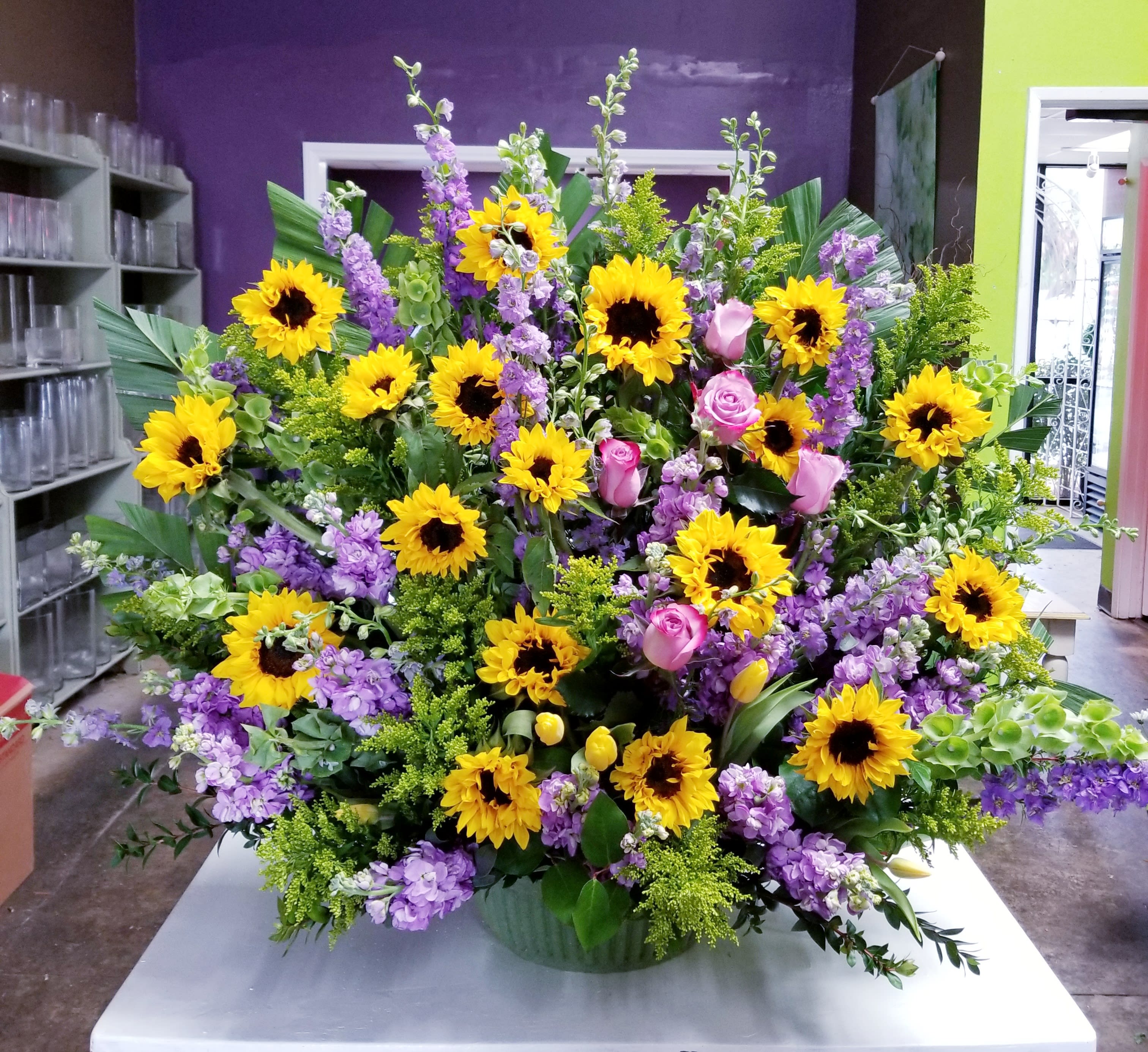Large arrangement of yellow sunflowers, pink roses, and purple flowers in a green container