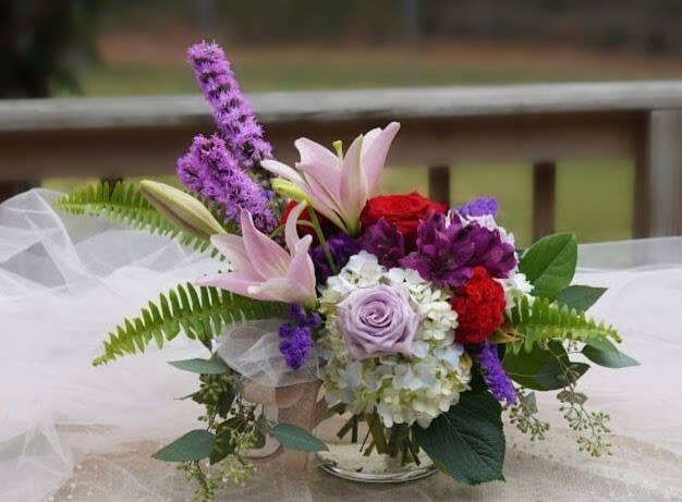 Presley's Passion - A garden-style cascade of pink lilies and lavender roses makes an immediate, romantic impression-perfect as an anniversary bouquet, Mother's Day gift, or a "just because" surprise for someone you adore. Presley's Passion is a flower arrangement designed to make the recipient feel truly seen: soft pastels meet vivid purple and red accents for a look that's both tender and dramatic, sending a clear message of love, celebration, or thoughtful sympathy depending on the moment.  This arrangement pairs fragrant pink lilies with creamy white hydrangea, lavender and red roses, bright alstroemeria, and vivid purple fillers for depth and texture. Displayed in a clear glass vase and finished with airy tulle wrap and long fern fronds, the loose asymmetrical design gives it a fresh, garden-picked movement-ideal for birthdays, get well wishes, new baby celebrations, thank yous, or springtime décor. Size and stem count vary by availability; water upon arrival and trim stems slightly to help blooms open and last longer.
