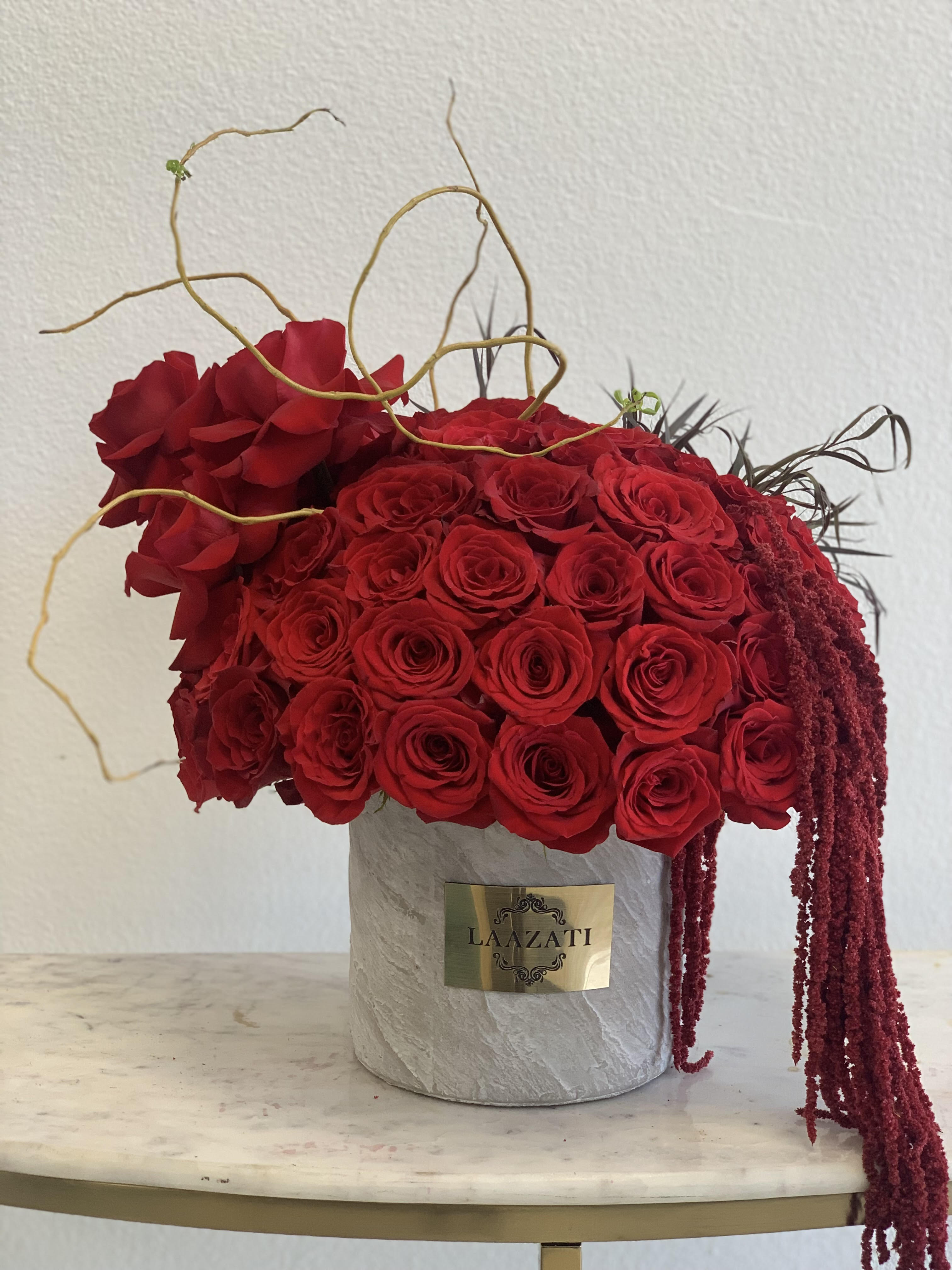 Red rose arrangement in a gray cylindrical box with trailing burgundy accents