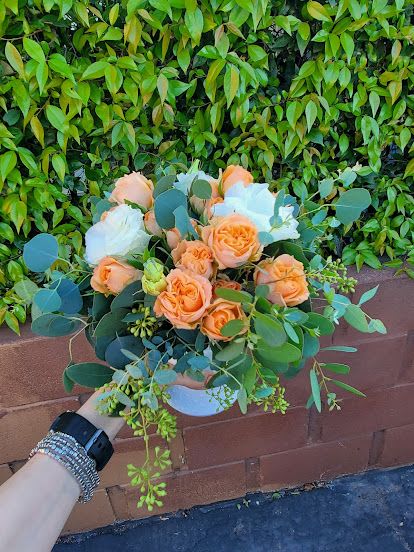 Low arrangement of peach roses and white blooms in a white bowl vase, held in front of a leafy hedge