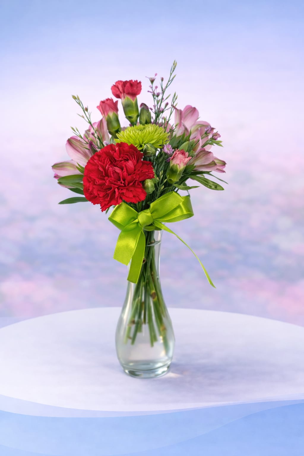 Sweetheart Pop - Bright, playful, and full of charm—Sweetheart Pop features bold red carnations paired with soft pink alstroemeria and a fresh pop of green button mums. Accented with delicate filler flowers and tied with a cheerful green ribbon, this petite arrangement is perfect for sending a little love in a big way.  Designed in a classic glass vase, it’s a sweet and simple choice for birthdays, thank-yous, or just because—proof that even the smallest bouquets can make the biggest impact. 