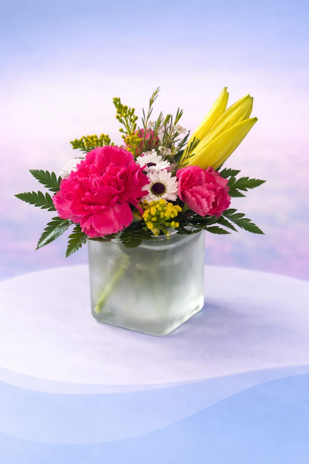 Sunshine Kiss - Bright and cheerful with a playful twist, Sunshine Kiss features vibrant pink carnations paired with sunny yellow lilies and delicate daisy accents. Designed in a modern frosted cube vase, this arrangement brings together bold color and soft texture for a look that’s both fun and fresh.  Perfect for lifting spirits, celebrating small moments, or adding a pop of happiness to any space, this design is a sweet reminder that a little sunshine goes a long way—Love Delivered. 