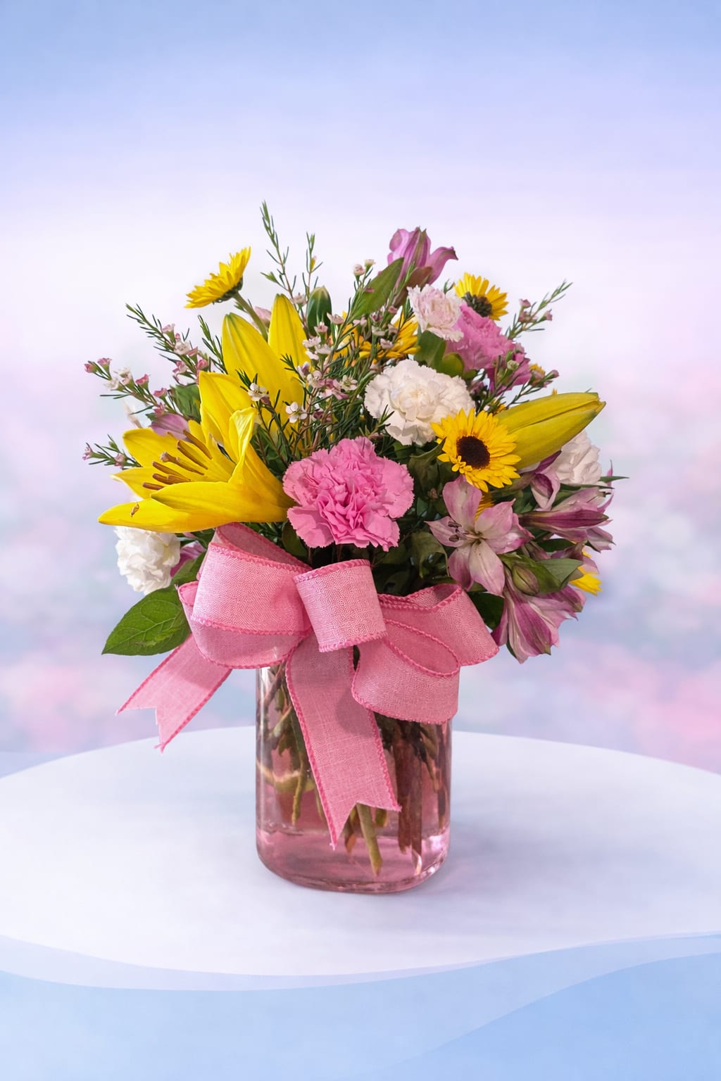 Blushing Sunshine - Bright, happy, and full of charm—Blushing Sunshine combines sunny yellow lilies and cheerful daisies with soft pink carnations and delicate blooms, all gathered in a blush-toned glass vase and finished with a sweet pink bow. This design brings together warmth and softness for the perfect balance of vibrant and romantic.  Ideal for birthdays, celebrations, or simply brightening someone’s day, this arrangement is a joyful burst of color designed to make them smile—Love Delivered. 