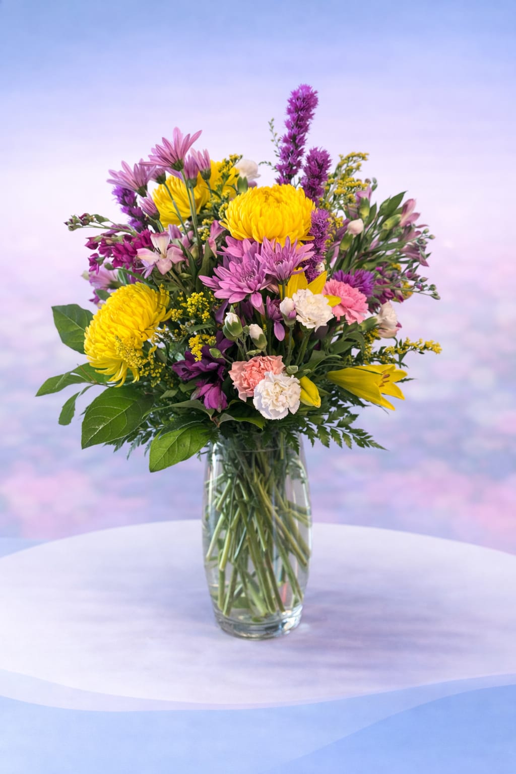 Wildflower Jubilee - Bright, joyful, and full of movement—Wildflower Jubilee is a lively mix of sunny yellow mums, playful purples, soft pinks, and delicate accents that feel freshly gathered from a blooming field. Designed in a classic clear vase, this arrangement has a natural, airy style with layers of texture and color that catch the eye from every angle.