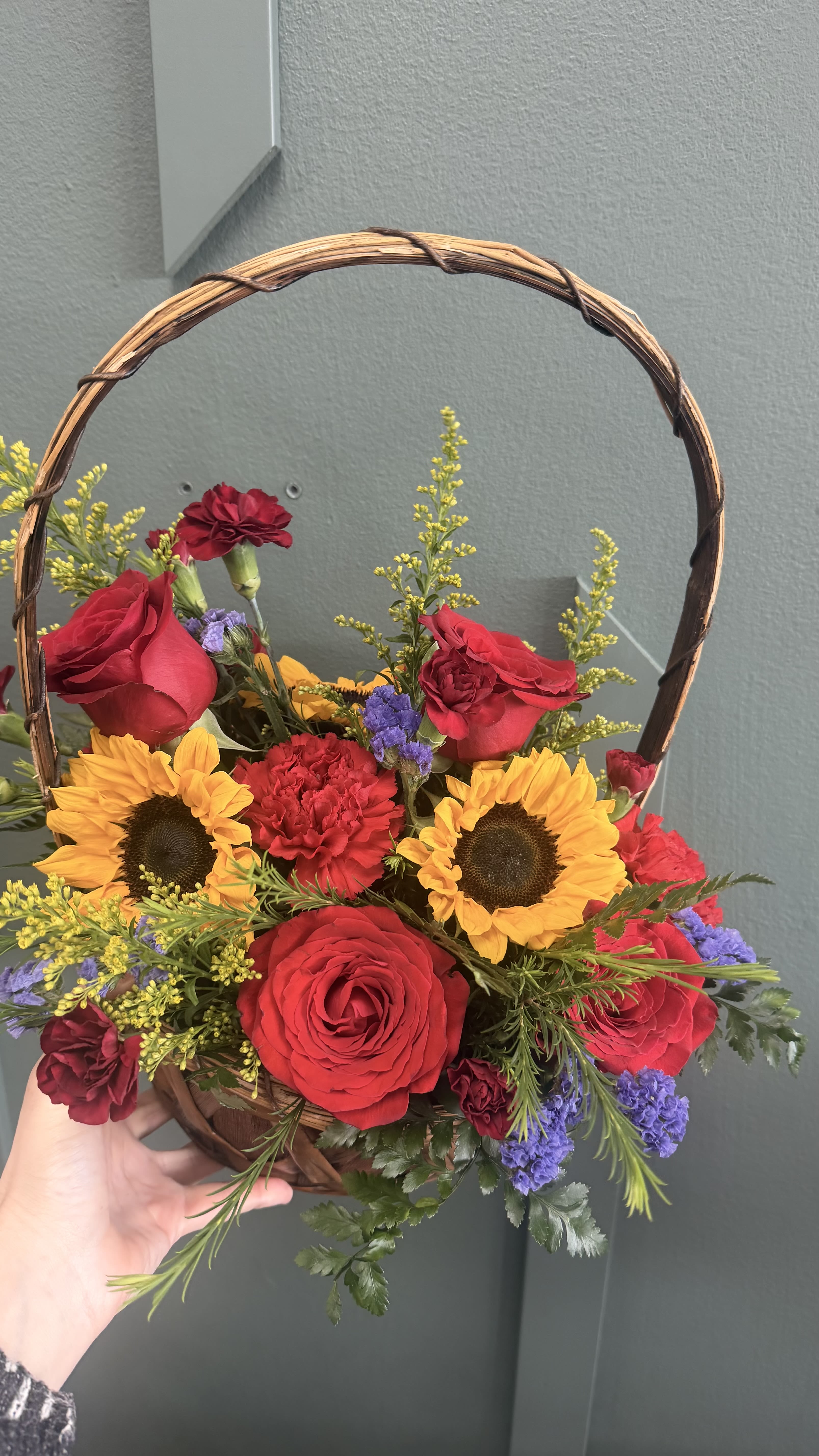 Roses and Sunflower basket - A bright, handpicked basket arrangement that pairs golden sunflowers with classic red roses and red carnations-perfect as a birthday bouquet, a cheerful get‑well gift, or a romantic surprise. The front-facing, rounded design and curved woven-handle basket give it a handcrafted, country-chic look that reads warm and joyful for Mother's Day, anniversaries, or simply to say "thinking of you."  This mixed-color arrangement adds depth with small purple filler blooms, fine-textured yellow accents, and feathery greenery for movement and contrast. Dense and abundant yet airy at the edges, the composition feels lively on a mantel, entry table, or beside a hospital bed. Available in a woven basket with a full, rounded face-sized to impress without overwhelming the room-and crafted to arrive fresh and ready to display.