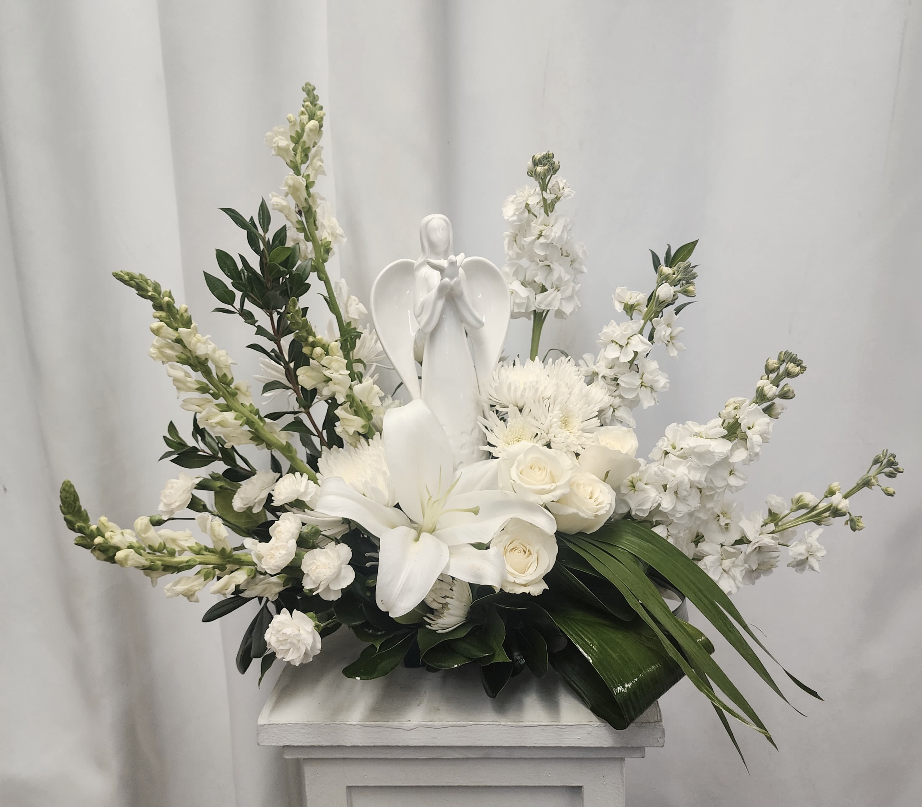 Angel of Passage - A peaceful arrangement of white lilies, roses, snapdragons, stock, cushions and mini carnations surrounds a classic keepsake ceramic angel with a bird in hand.  Lush green foliage adds contrast, while the overall composition evokes comfort and quiet remembrance — a graceful tribute for moments of reflection. 