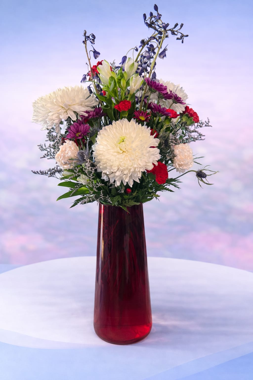 Crimson Elegance - Striking and sophisticated, Crimson Elegance features lush white spider mums and soft cream blooms accented with rich red carnations and touches of deep plum and airy textures. Designed in a bold ruby glass vase, this arrangement brings height, movement, and contrast for a look that feels both dramatic and refined.