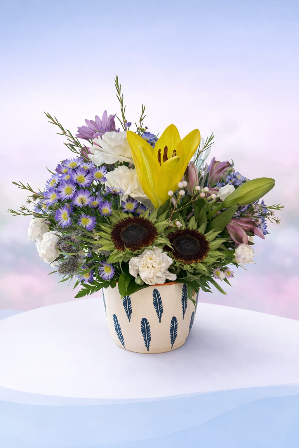 Sunny Meadow Song - Bright, cheerful, and full of personality—Sunny Meadow Song features radiant yellow lilies and sunny sunflowers nestled among soft lavender blooms, crisp white carnations, and delicate wildflower accents. Designed in a charming patterned container, this arrangement has a fresh-from-the-meadow feel with a playful, uplifting spirit.