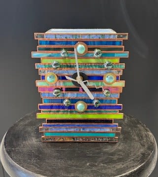 #A136 - Striped Clock - A playful, modern decorative clock arranged like a small art object makes a cheerful gift for birthdays or housewarmings - the kind of accent that makes the person you're thinking of smile every time they pass it. This clock is made with mixed-colored glass accents that create a bright, contemporary presentation suited for casual celebrations or someone with an eclectic, colorful home. Think of it as a practical keepsake that reads like a mini sculpture.  The design features a stacked rectangular face of horizontal bands in blue, purple, pink, yellow and turquoise tones, sitting on a dark pedestal base, that lets the vivid colors pop. A companion rectangular decorative box echoes the same layered-strip motif in blue, teal, green, peach and purple, detailed with bead-like accents and a central circular ornament for handcrafted texture. Ribbon and beads complete the composition, giving the arrangement a tactile, gift-ready finish.  Perfect for a friend's new apartment, a cheerful birthday surprise, or a casual "thinking of you" present, this piece arrives ready to display on a mantel, bookshelf, or bedside table where its bright palette will lift the room. Available in a single size; minimal care required since the focal elements are decorative hard goods - simply dust occasionally and enjoy the long-lasting color and charm.