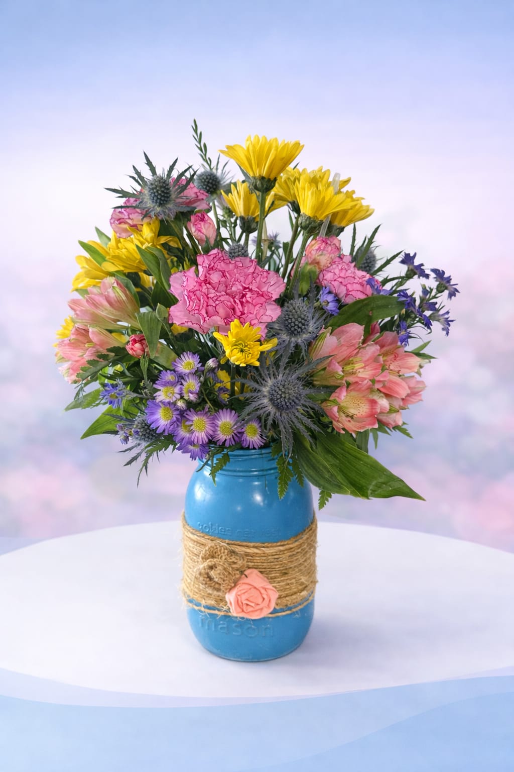 Rustic Wildflower Breeze - Colorful, textured, and full of natural charm—Rustic Wildflower Breeze features a lively mix of sunny yellow daisies, soft pink carnations, delicate lavender accents, and unique thistle for a touch of wild beauty. Designed in a painted mason jar wrapped with rustic twine, this arrangement has a relaxed, countryside feel with just the right amount of personality.