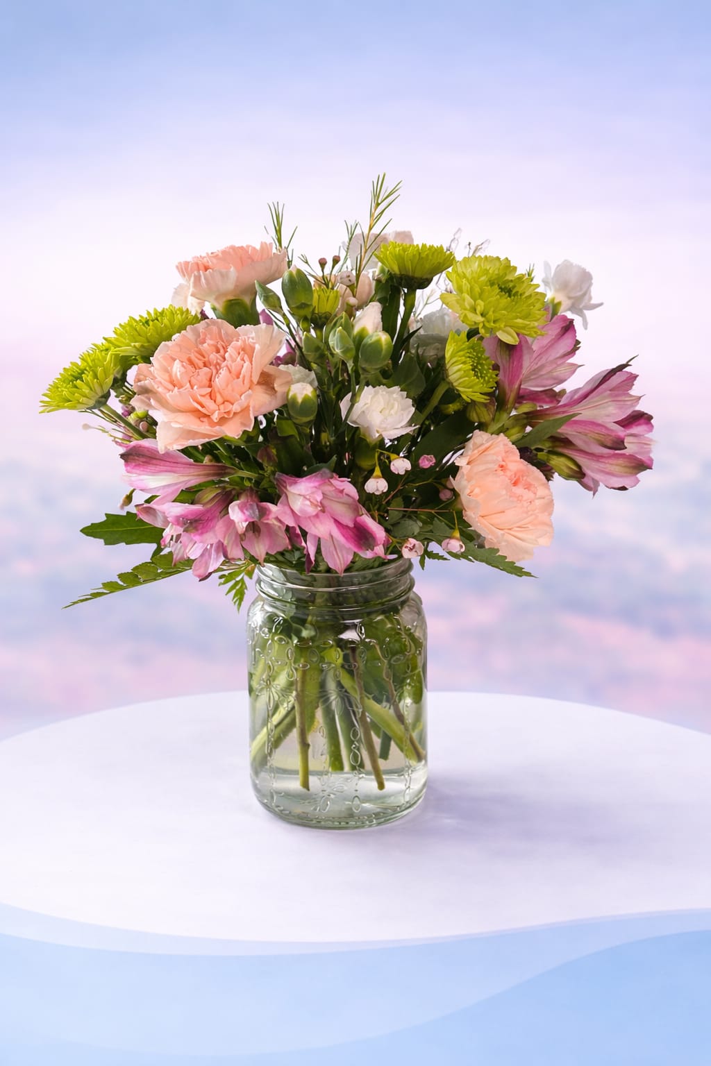 Blush Garden Glow - Soft peach carnations and delicate pink alstroemeria come together with fresh green button mums and airy white accents in this light, cheerful design. Arranged in a classic glass jar, Blush Garden Glow has a fresh-picked, garden-inspired feel that’s both sweet and uplifting. Perfect for everyday moments, thank-yous, or a simple “thinking of you,” this arrangement brings a gentle pop of color and a whole lot of heart—beautifully designed to deliver love. 
