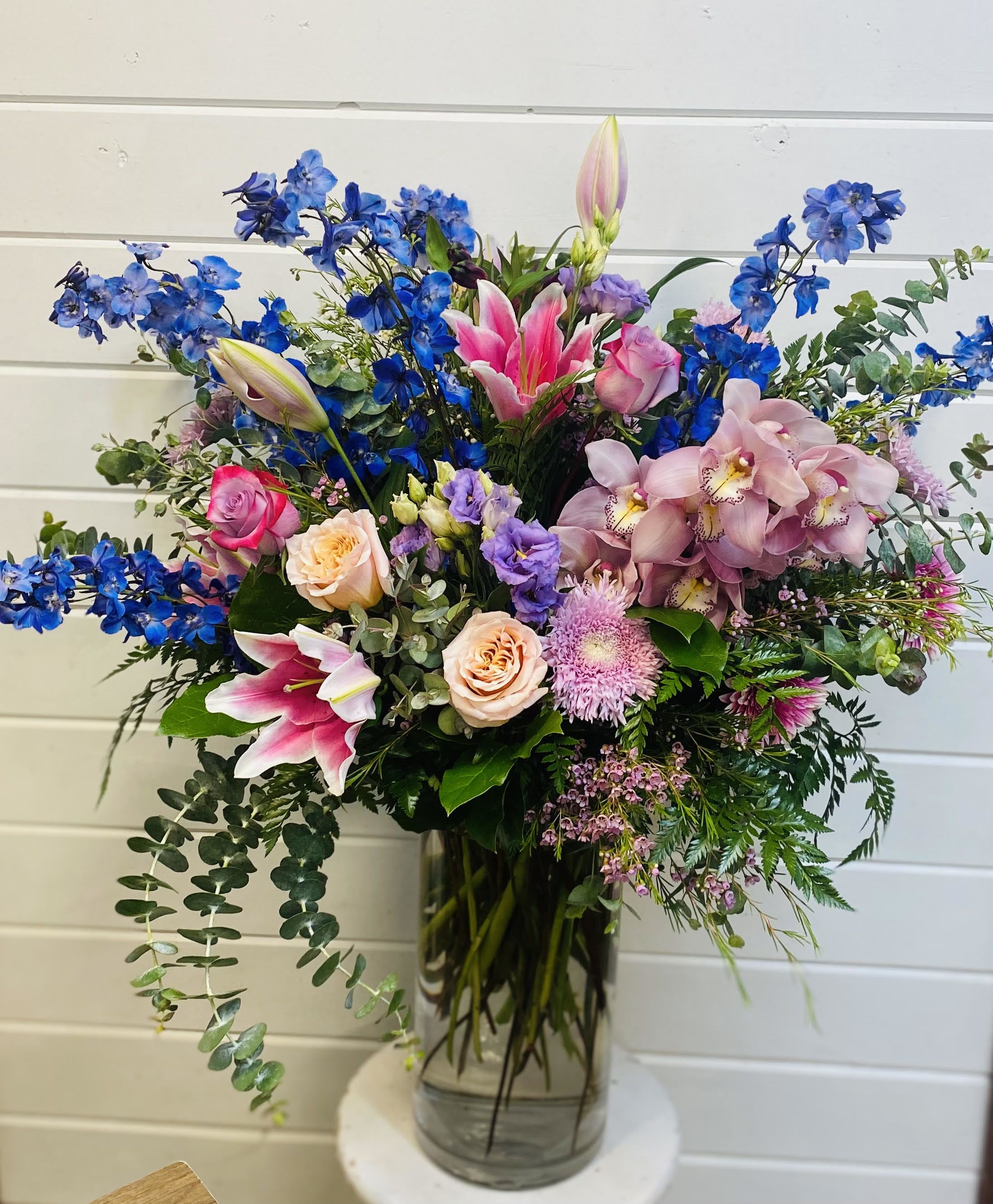 Tall mixed arrangement of pink lilies, roses, orchids and blue delphinium in a clear glass vase
