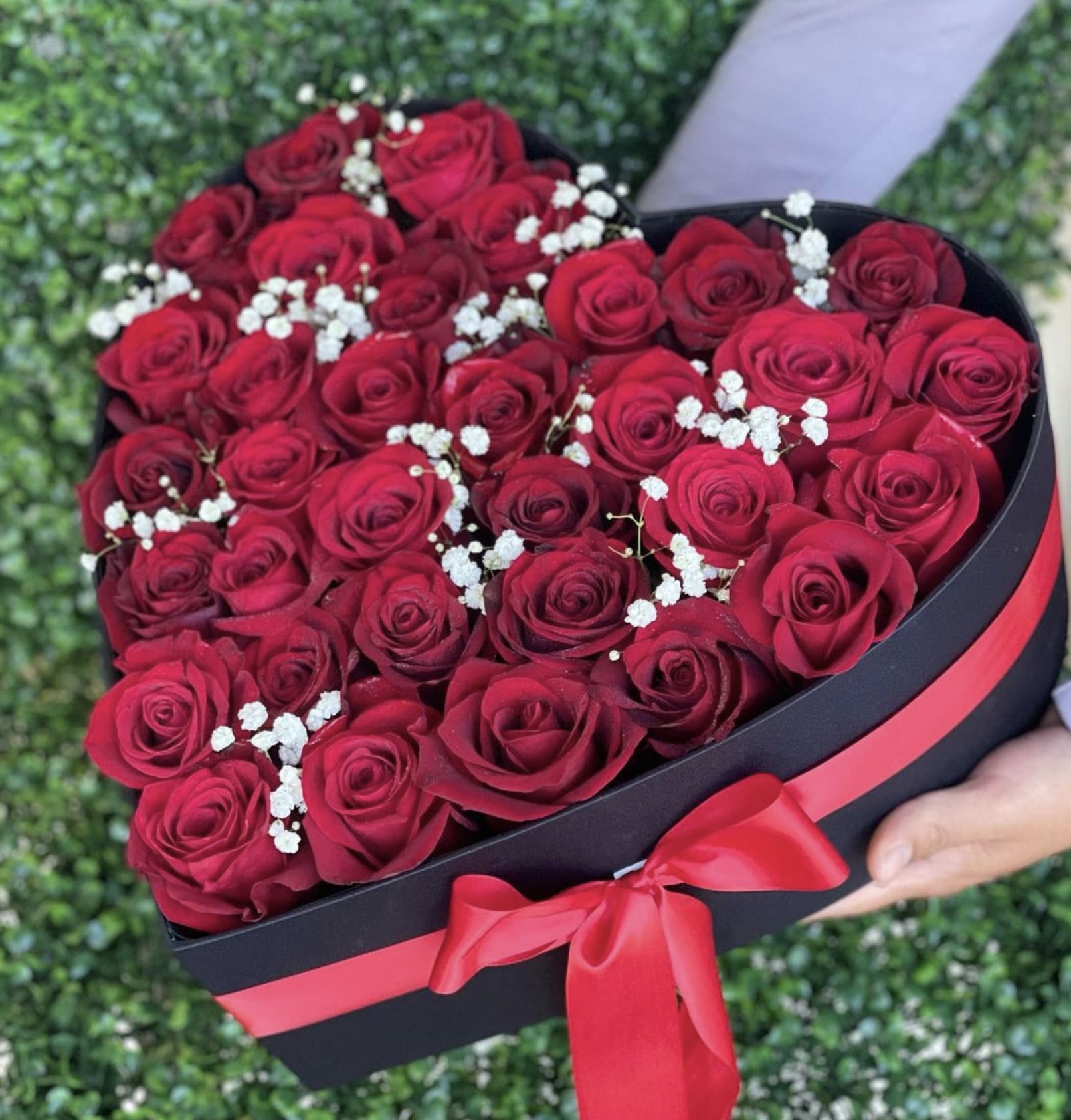Heart-shaped black box filled with red roses and small white blooms, tied with a red ribbon.