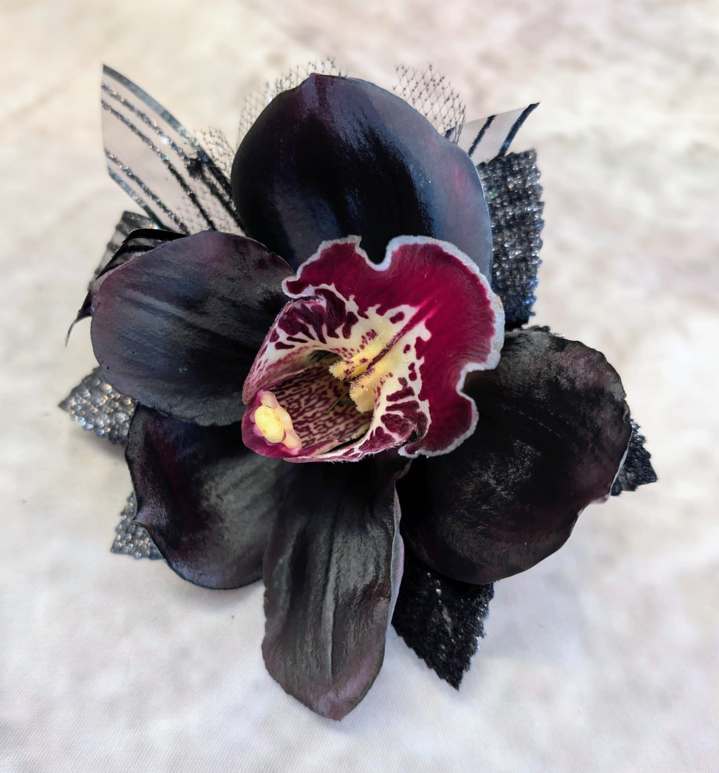 Noir Orchid Boutonniere - Dark, dramatic, and undeniably refined, this black orchid boutonnière is a statement in modern elegance. Featuring a velvety deep-toned cymbidium orchid with rich plum undertones and a striking wine-colored center, this piece evokes a sense of mystery and quiet luxury. Subtle accents of textured ribbon and shimmering black details add depth and a couture-inspired finish.  Handcrafted at Windmill Floral Expressions in Las Vegas, each boutonnière is designed with intention—perfect for weddings, proms, black-tie events, or anyone drawn to a bold, unconventional floral aesthetic.  This piece pairs beautifully with formalwear, offering a sophisticated alternative to traditional florals. As with all of our designs, each boutonnière is custom made, ensuring a one-of-a-kind creation tailored to your event.  Available for same-day flower delivery in Las Vegas and Henderson, or curbside pickup by appointment.