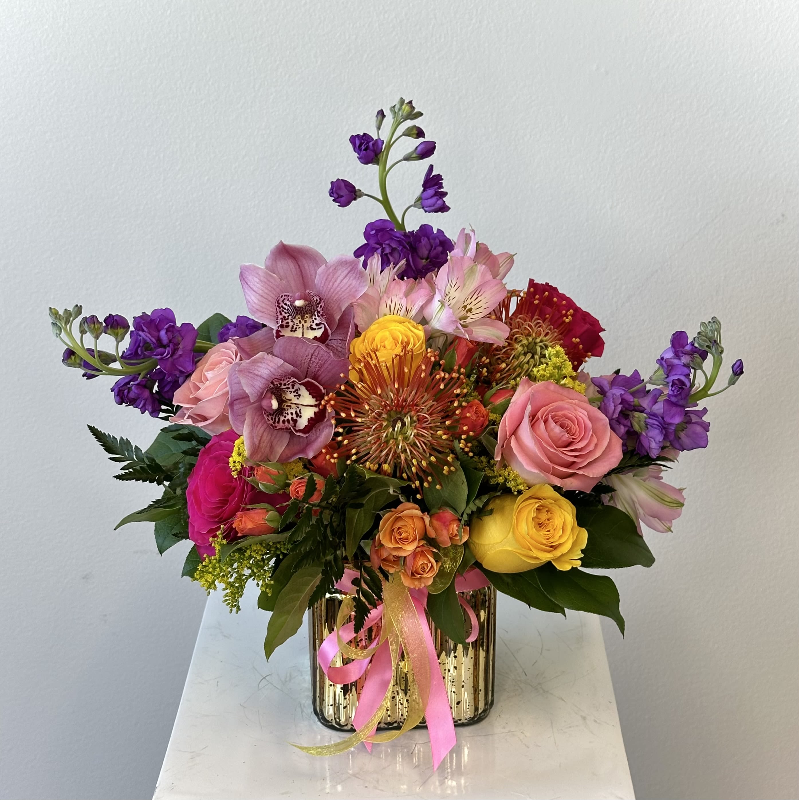 Colorful bouquet of roses, orchids, and mixed blooms in a gold vase with pink ribbon