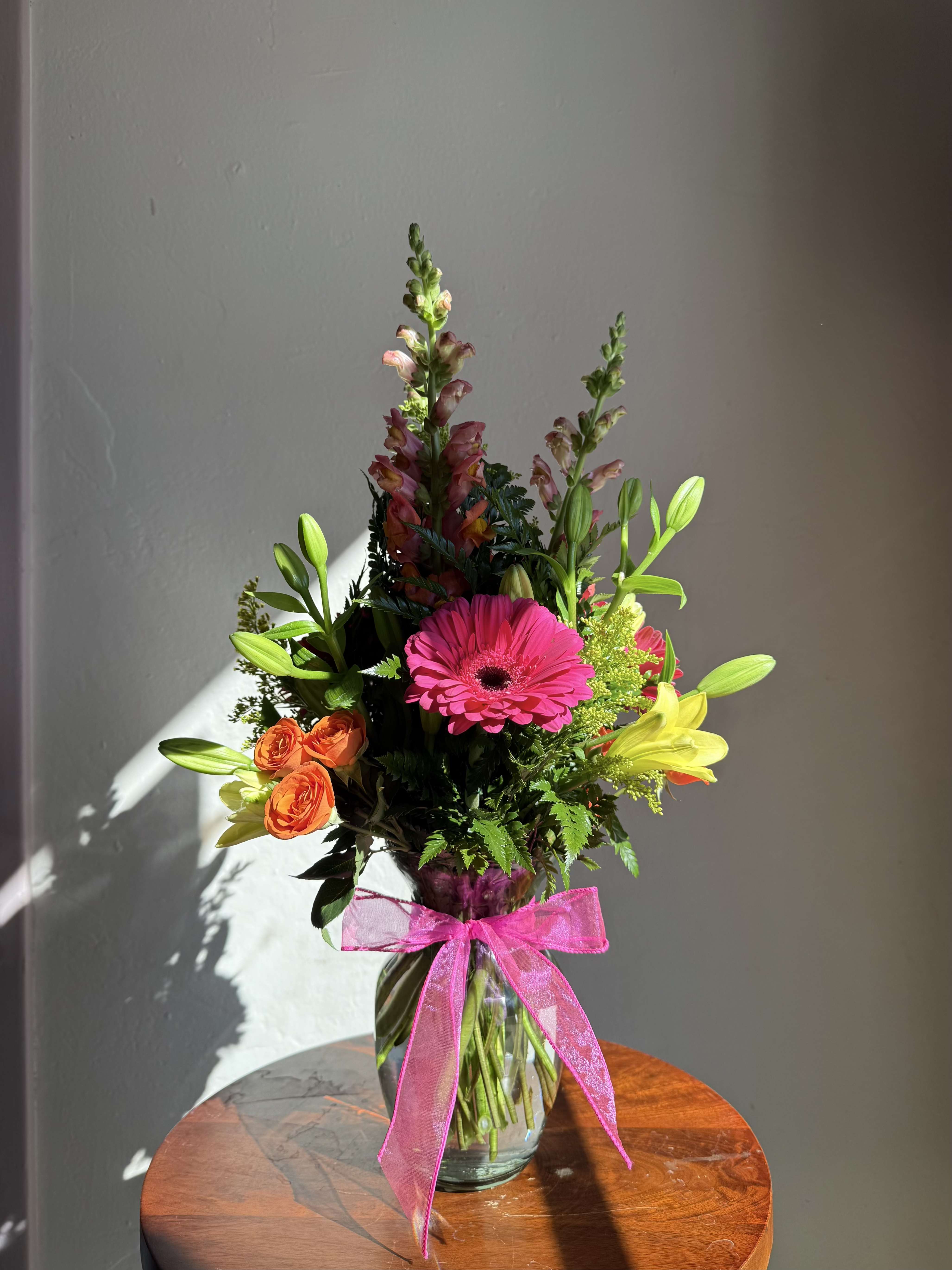 Tall mixed bouquet with pink gerbera daisy, orange roses, lilies and snapdragons in a clear vase with pink bow