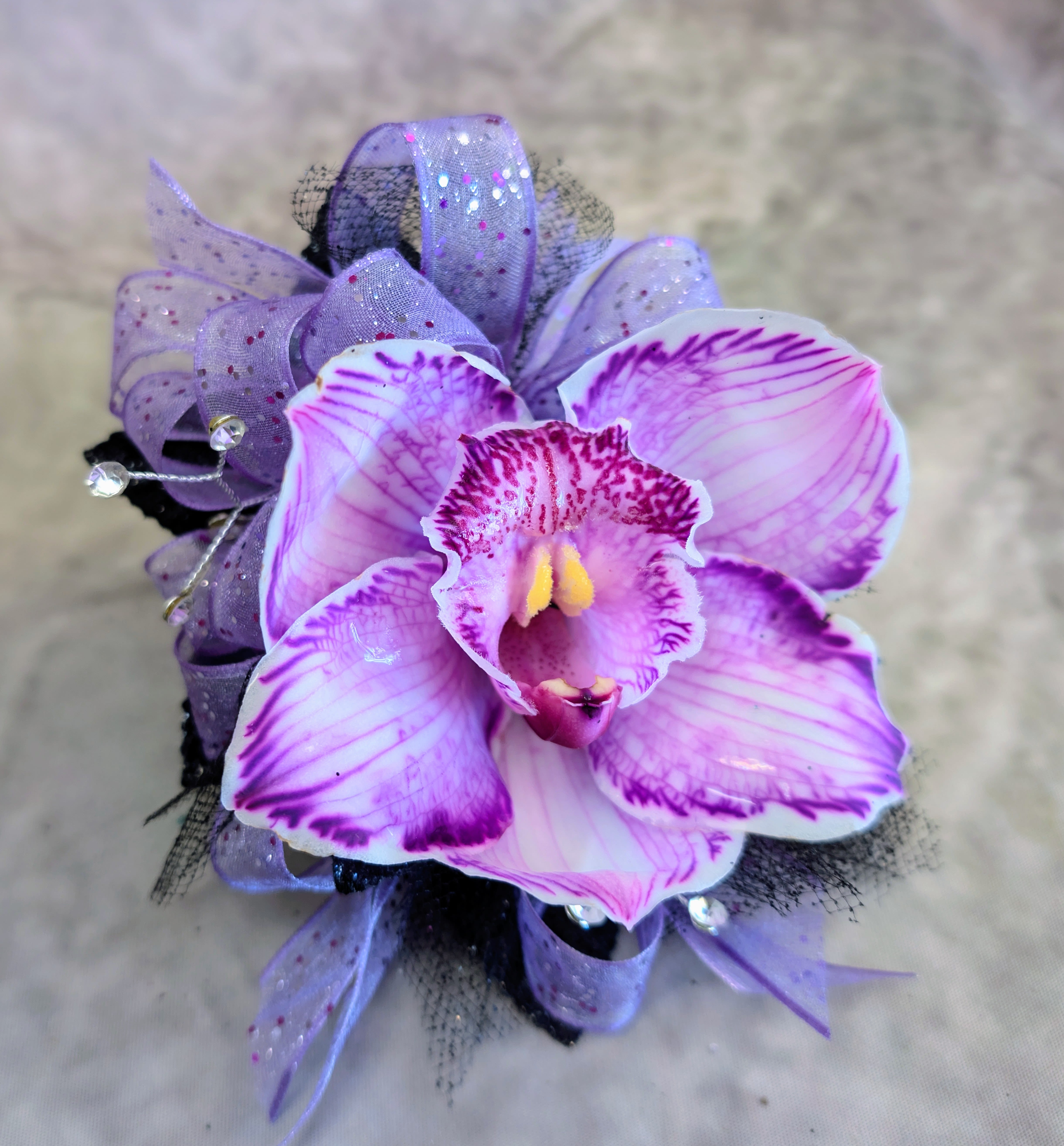 Violet Orchid Corsage - Soft, luminous, and effortlessly enchanting, this corsage features a radiant lavender cymbidium orchid kissed with vivid magenta veining and a glowing golden center. The bloom is framed with airy layers of sheer ribbon, delicate tulle, and subtle crystal accents that catch the light with every movement, creating a dreamy, ethereal finish.  Handcrafted at Windmill Floral Expressions in Las Vegas, this corsage blends romance with a touch of modern sparkle—perfect for proms, weddings, or any moment that calls for something truly special. Each piece is custom designed, ensuring no two are ever exactly alike. 