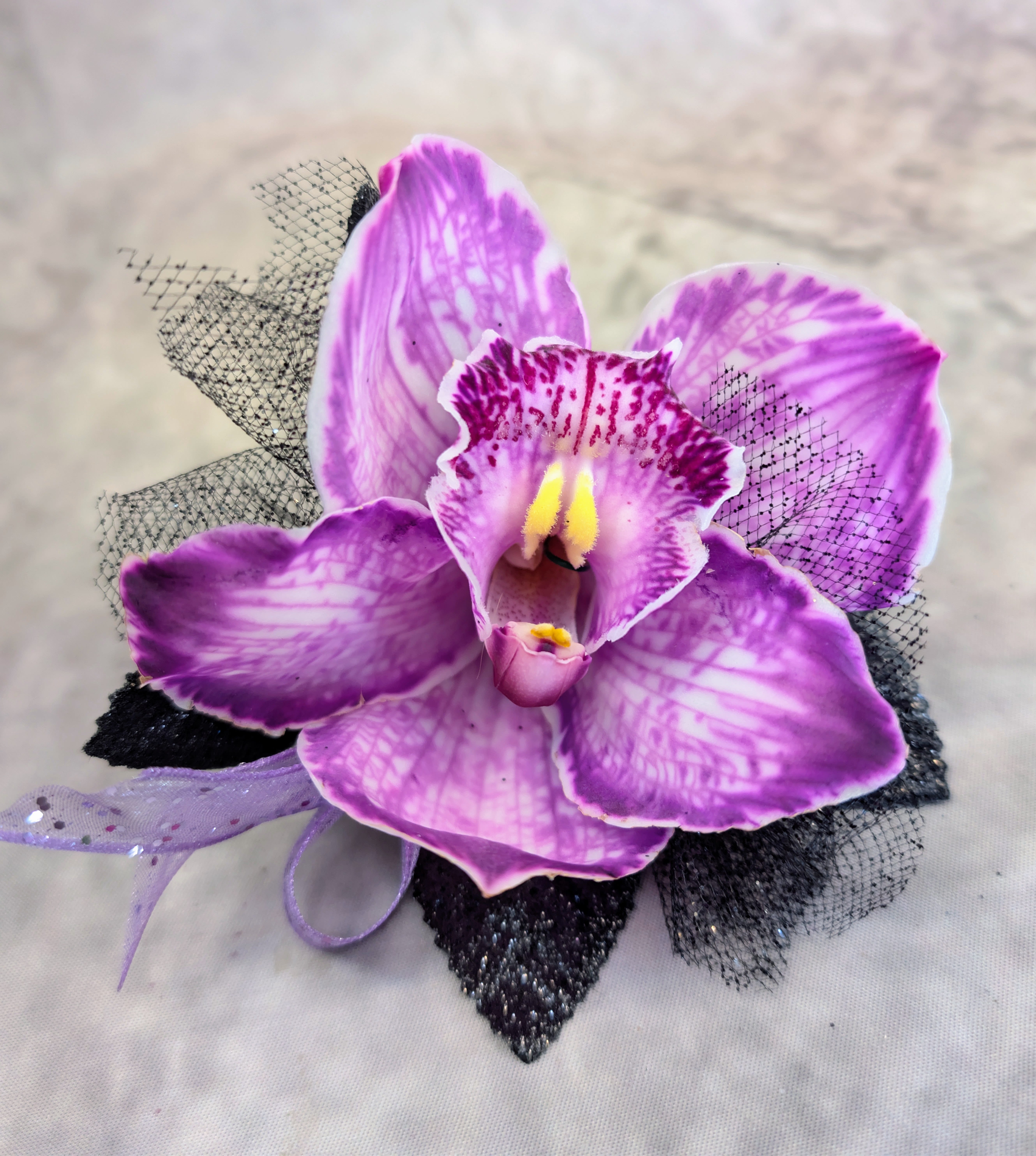 Lavender Orchid Boutonniere - Soft yet striking, this lavender cymbidium orchid boutonnière features delicate magenta veining and a luminous golden center, creating a beautifully dimensional bloom. Accented with airy black tulle, subtle shimmer, and flowing ribbon details, it offers a perfect balance of romance and modern edge.  Handcrafted at Windmill Floral Expressions in Las Vegas, each boutonnière is thoughtfully designed to complement weddings, proms, and special occasions with a touch of elevated elegance. No two pieces are exactly alike, ensuring a custom, one-of-a-kind finish.