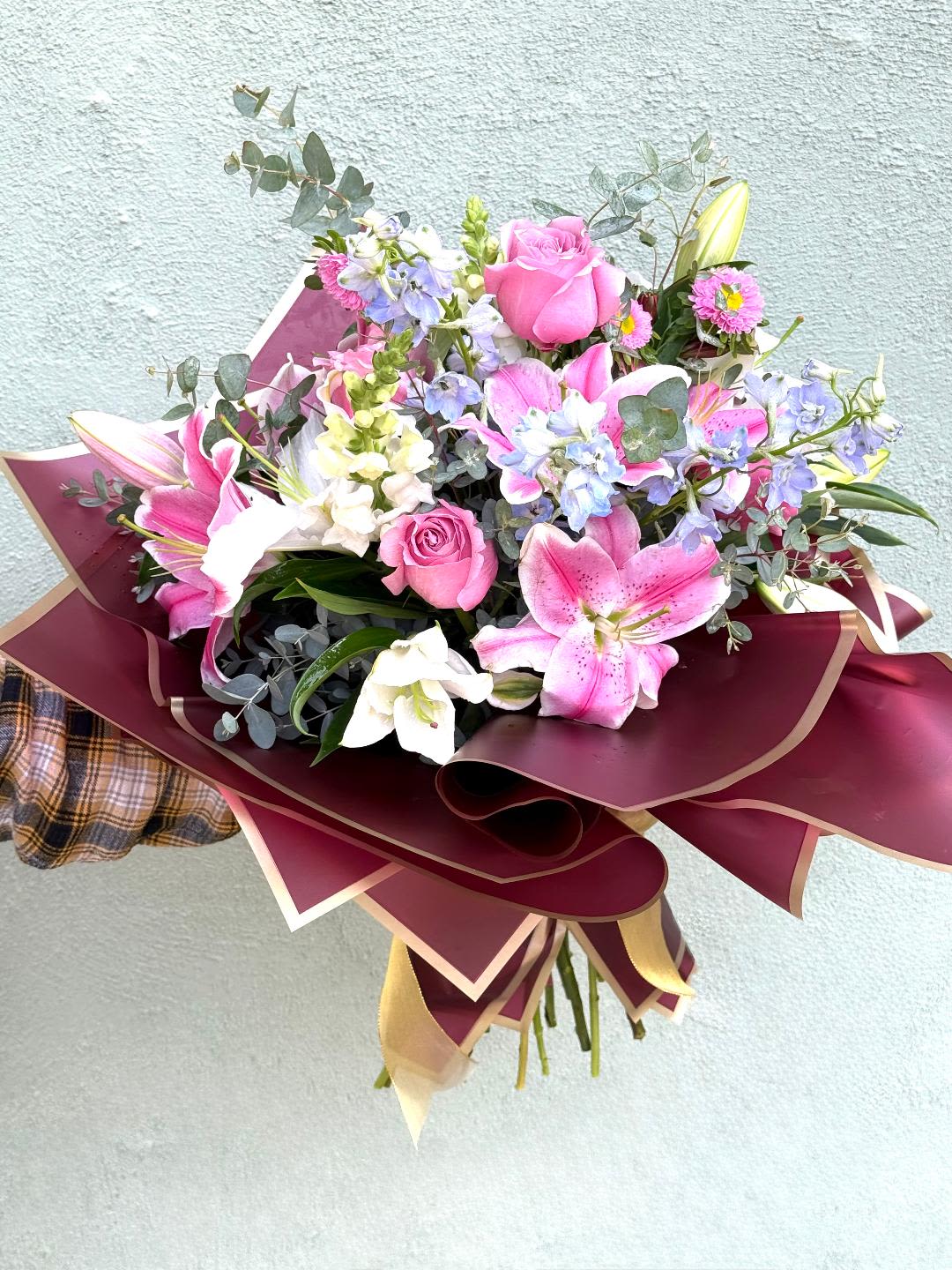 Hand-tied bouquet of pink roses, lilies, and mixed blooms wrapped in burgundy and gold paper.