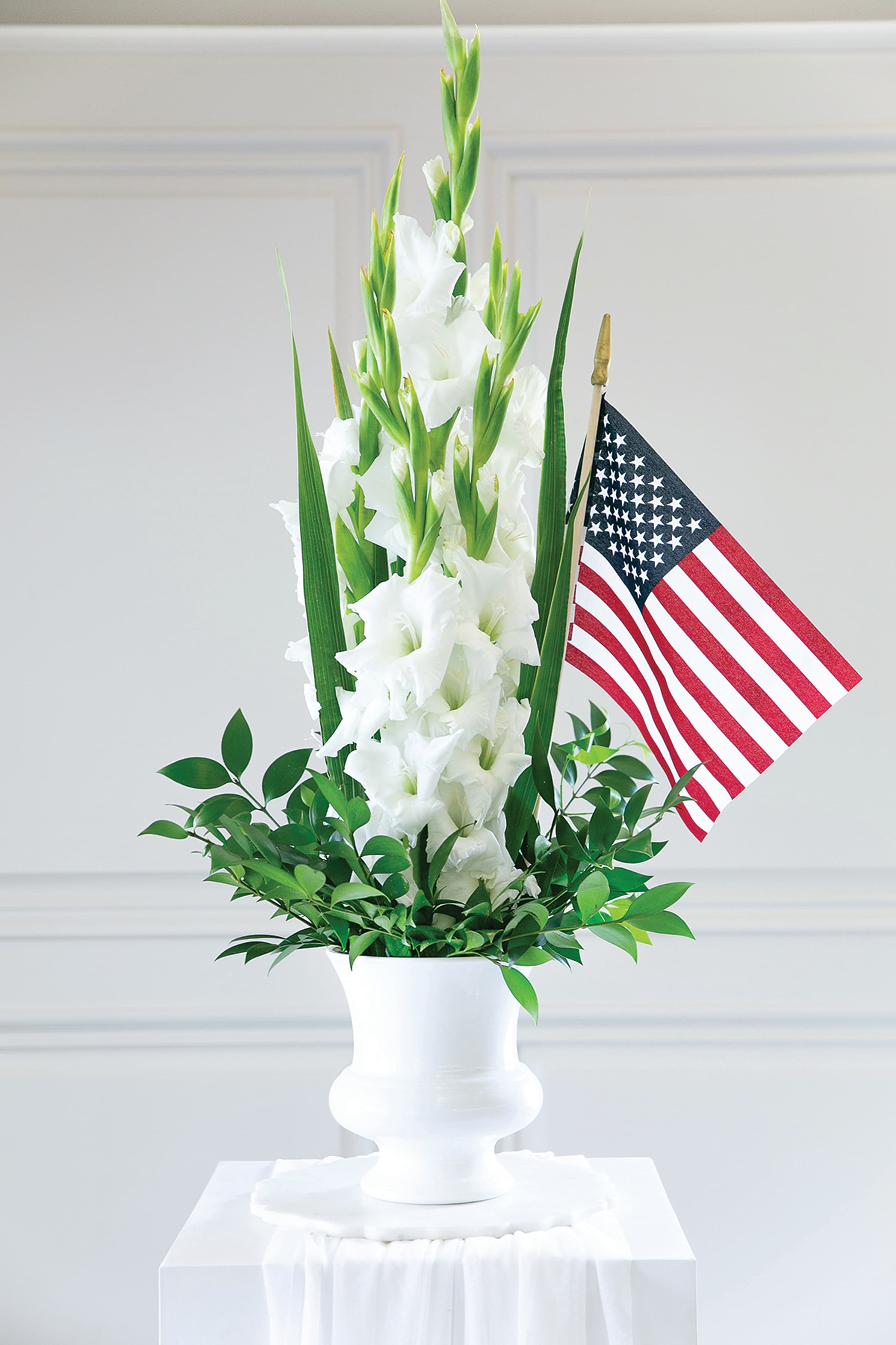 White gladiolus arrangement in a white urn with an American flag