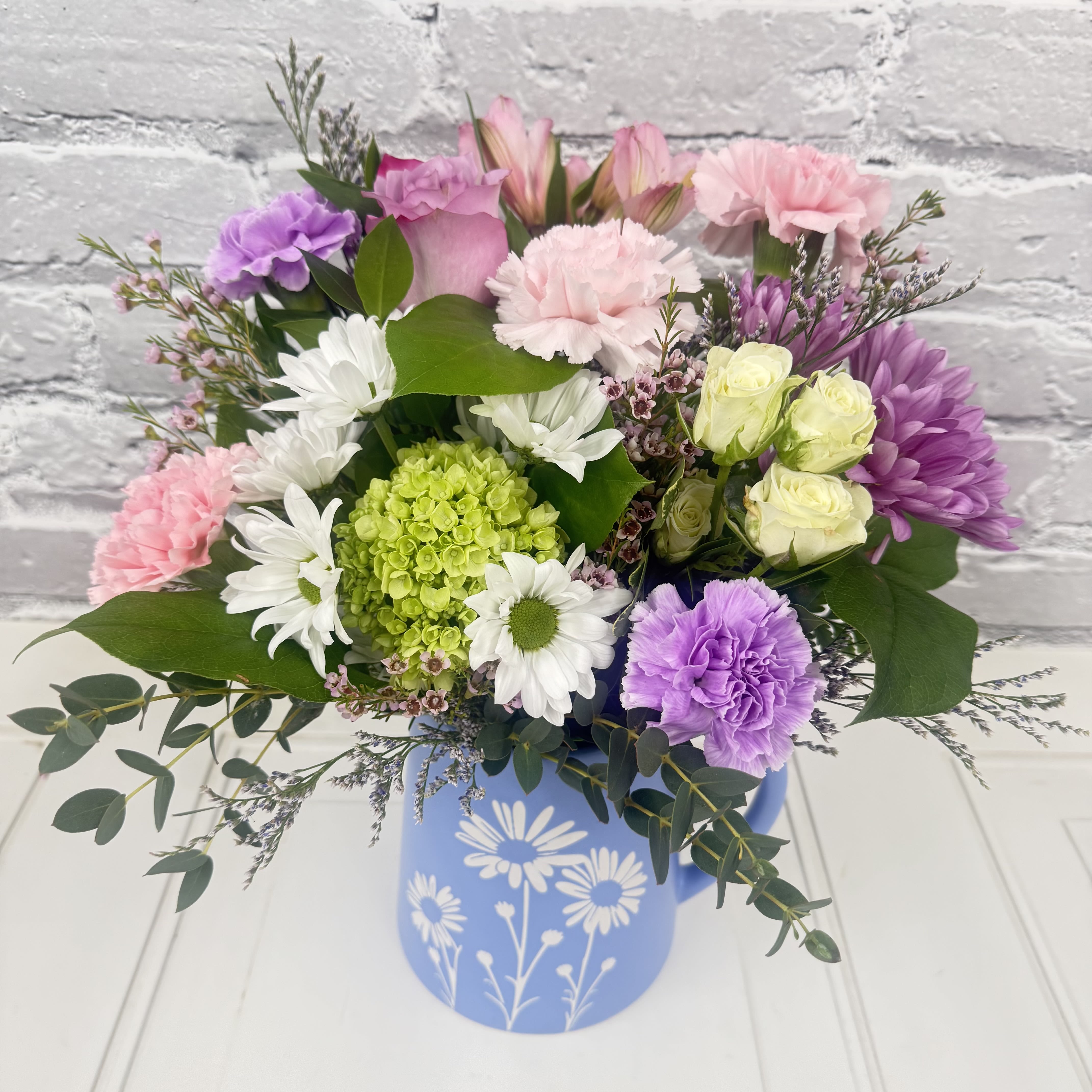 Mom's Daisy Pitcher - Soft pink carnations and lavender chrysanthemums mingle with pale roses and white daisies in a garden-fresh arrangement made for Mother's Day, birthdays, or "just because" moments. The Mom's Day Daisy Pitcher arrangement presents a bright green hydrangea focal point and touches of blue and green foliage that make the bouquet feel airy and layered-perfect for the person you want to brighten with a cheerful, homey surprise.  Arranged in a matte blue ceramic pitcher decorated with white daisy-style graphics, the mix includes daisies, alstroemeria, hydrangea, roses, carnations and complementary filler for rounded texture and visual depth. The casual pitcher serves as both vase and keepsake décor, bringing a soft palette of pink, purple, white, green and blue to tabletops, kitchen counters, or bedroom dressers.  This is the kind of gift that makes Mom feel seen and celebrated-a sweet, everyday statement for spring afternoons, thank-you moments, or a birthday brunch centerpiece. Pitcher size and exact stem counts vary by freshness and seasonal availability; simple care is to trim stems, refresh water daily, and keep away from direct heat to extend the bloom life.