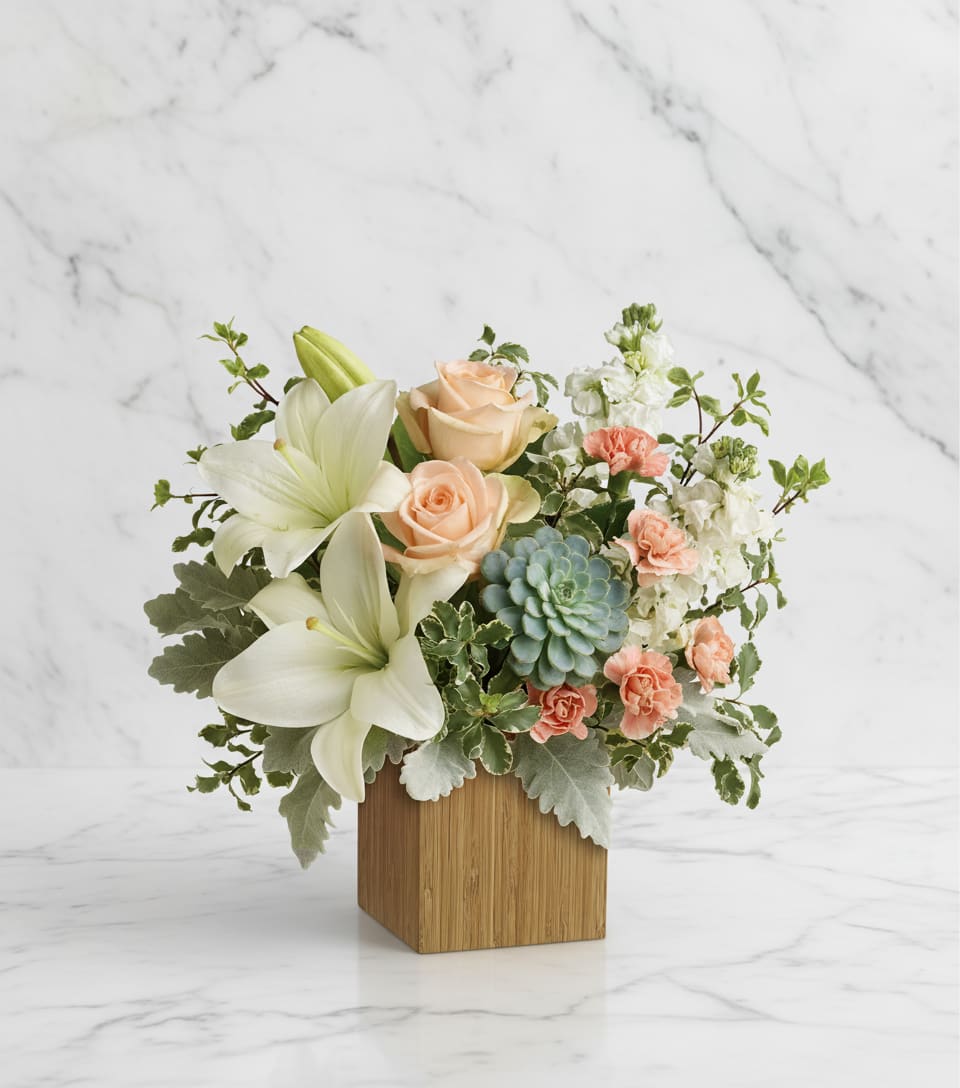 Desert Sunrise Box - Peaceful yet energizing, this unforgettable arrangement of desert-hued blooms and succulents in a sleek bamboo cube is a chic gift on any special occasion. Peach roses, white asiatic lilies, peach miniature carnations, and white stock are accented with pitta negra, dusty miller, and a green echeveria succulent. Delivered in a Small wood cube  Please Note: The bouquet pictured reflects our original design for this product. While we always try to follow the color palette, we may replace stems to deliver the freshest bouquet possible, and we may sometimes need to use a different vase.