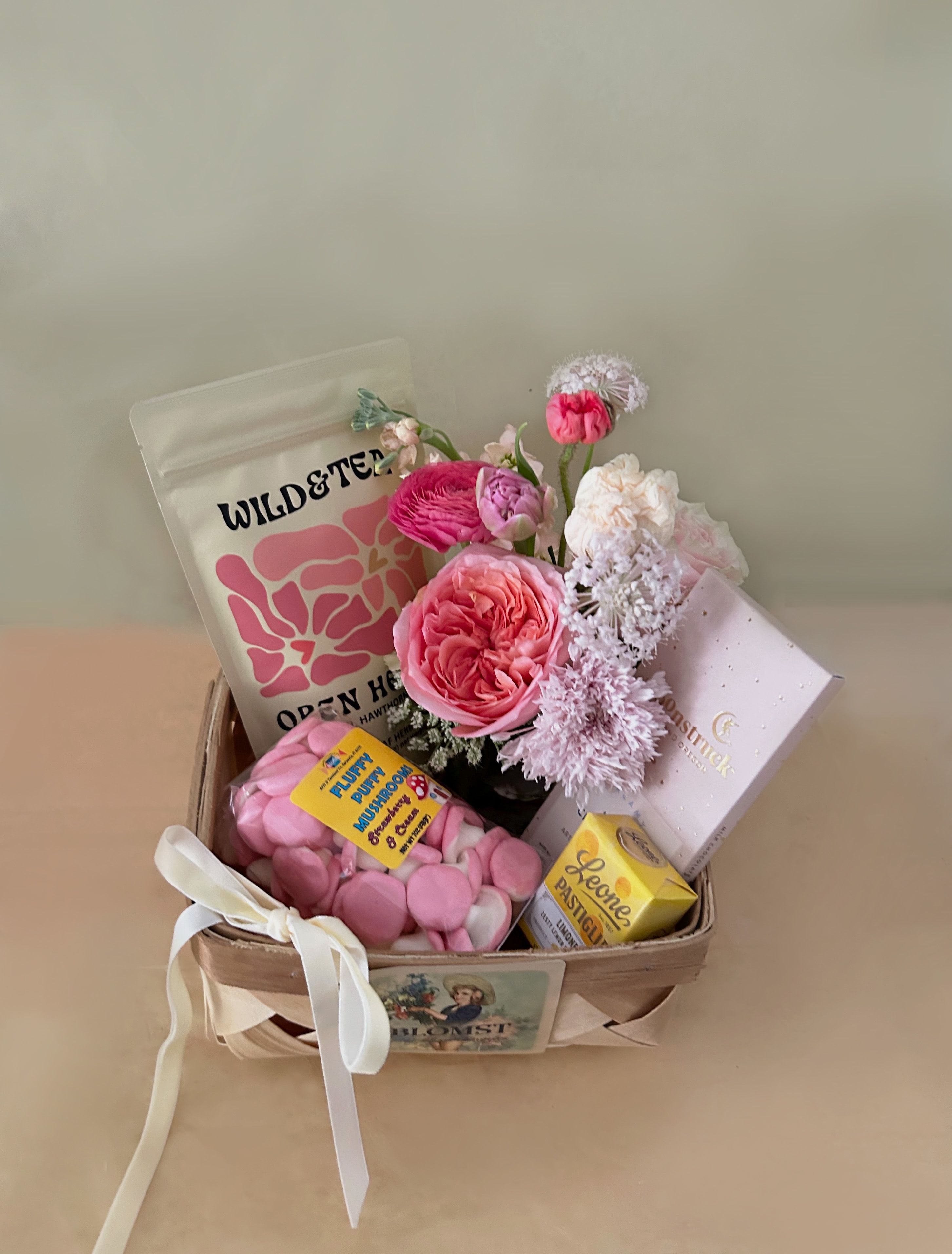 Carrie Candy Basket - A lovely, sweet  basket in the colors of light pink, and hot pink in the flavors of sweet candy and lemon. The petite and classic wooden French basket measures 7" by 7" by 4" and is overflowing with lovely treats and flowers. The basket contains: Wild &amp; Tea herbal tea "Open Heart": Divine self-love. Sweet, comforting and uplifting. Nurtures both physical and emotional heart health.  Organic Ingredients- Linden flower, hawthorn leaf and flower and roses. Caffeine free Moonstruck Creamy Milk chocolate bar: Everything is influenced by the gravitational pull of the moon, so it's no coincidence you were drawn to this creamy milk chocolate with a light touch of vanilla. Fluffy Puffy Mushrooms: Light, Swedish favorite mushroom shaped marshmallow. Strawberry and Cream Flavored. 7 ounces. Pastiglie Leone's Candy Originals are an Italian classic. These small delights are the result of a careful choice of essences, extracts and herbs. Their pleasant pastel shades (made from natural ingredients) make this product alluring to the eye as well as to the palate. A mini Blomst arrangement of fresh, pretty stems in a clear cylinder vase 4" by 4". Velvet riboon. ready for hand delivery in LA.