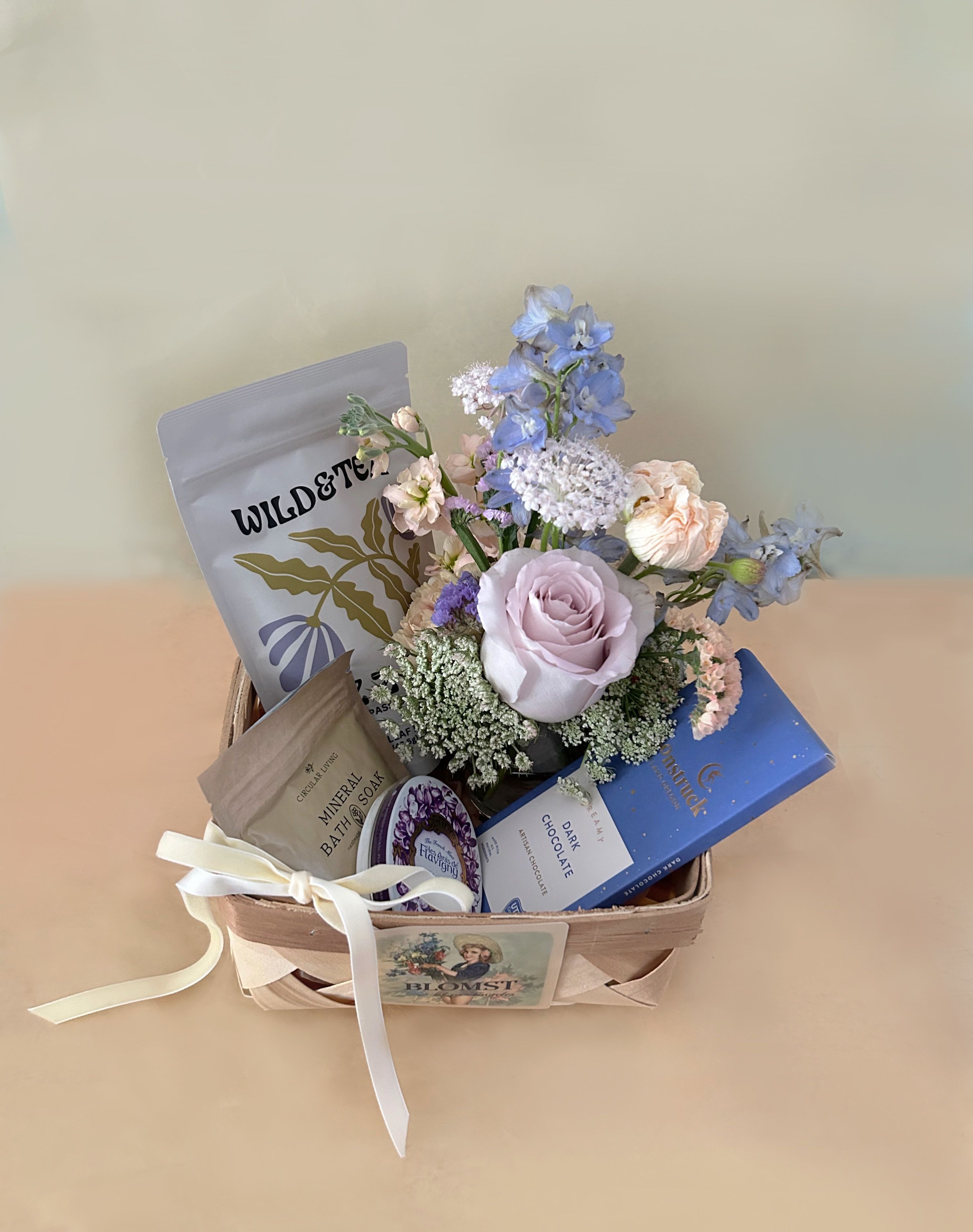Blue Velvet Basket - A classic and beautiful basket in the muted colors of light lavender, blue and white in the flavors of violet, cocoa and herbs. The petite and classic wooden French basket measures 7" by 7" by 4" and is overflowing with lovely treats and flowers. The basket contains: Wild &amp; Tea herbal tea "Zzzzzz": Smooth and minty, with subtle undertones of hops and lavender. Calm your nervous system and effortlessly drift into a peaceful sleep.  Organic Ingredients- Skullcap, chamomile, passionflower, peppermint, hops and lavender. Caffeine free Moonstruck Dreamy Dark Chocolate. Like a moonless sky, this rich chocolate captures a mesmerizing darkness. Let its deep roasted notes and curious hint of fruit light the way.  Circular living mineral bath soak. Eucalyptus/Green Tea natural bath salt to soothe and calm the skin. Les Anis De Flavigny: Floral and sweet, these Violet mints will leave you feeling minty and fresh.These all-natural violet flavored candies are housed in a vintage decorative tin, with about 30 mints per tin. A mini Blomst arrangement of fresh, pretty stems in a clear cylinder vase 4" by 4". Velvet ribbon. ready for hand delivery in LA.