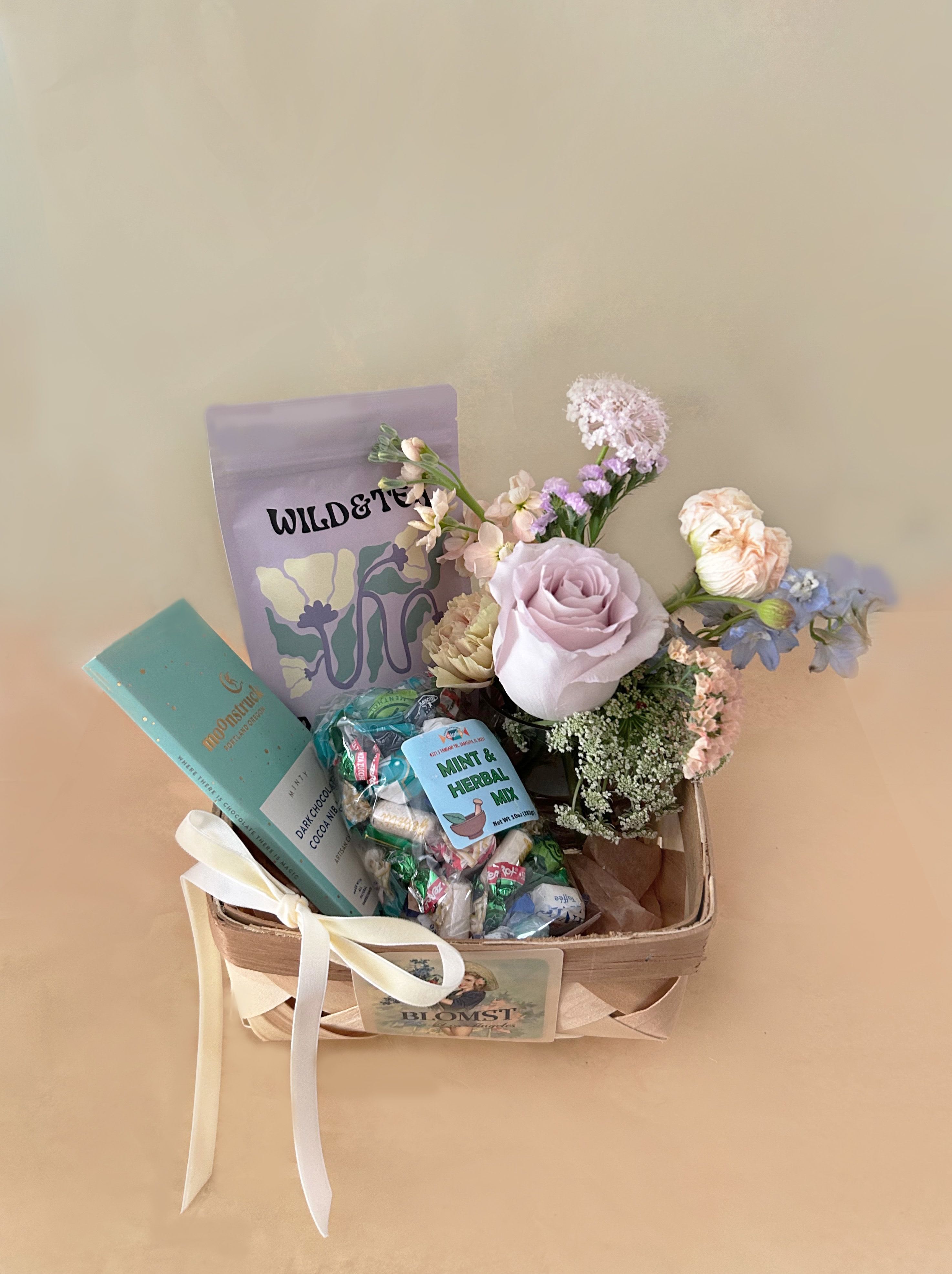 Mila Mint basket - Such a pretty basket in the colors of lavender and light turquoise in the flavors of sweet mint and herbs. The petite and classic wooden French basket measures 7" by 7" by 4" and is overflowing with lovely treats and flowers. The basket contains: Wild &amp; Tea herbal tea: Delicate floral aroma with a hint of sweetness. Unwind muscle tension and alleviate minor aches and pains, especially effective for headaches. Organic ingredients: meadowsweet, wood betony, chamomile, peppermint and lavender.  Moonstruck Minty Dark chocolate bar: Fresh notes of Oregon mint flourish in the dark chocolate depths of this elegant bar, studded with a constellation of cocoa nibs. Mint and Herbal Candy Mix 10oz Bag. The bag is filled with a variety of hard candies that bring together unique flavors. Indulge in a delightful mix of European mint and herbal candies A mini Blomst arrangement of fresh, pretty stems in a clear cylinder vase 4" by 4". Velvet riboon. ready for hand delivery in LA.
