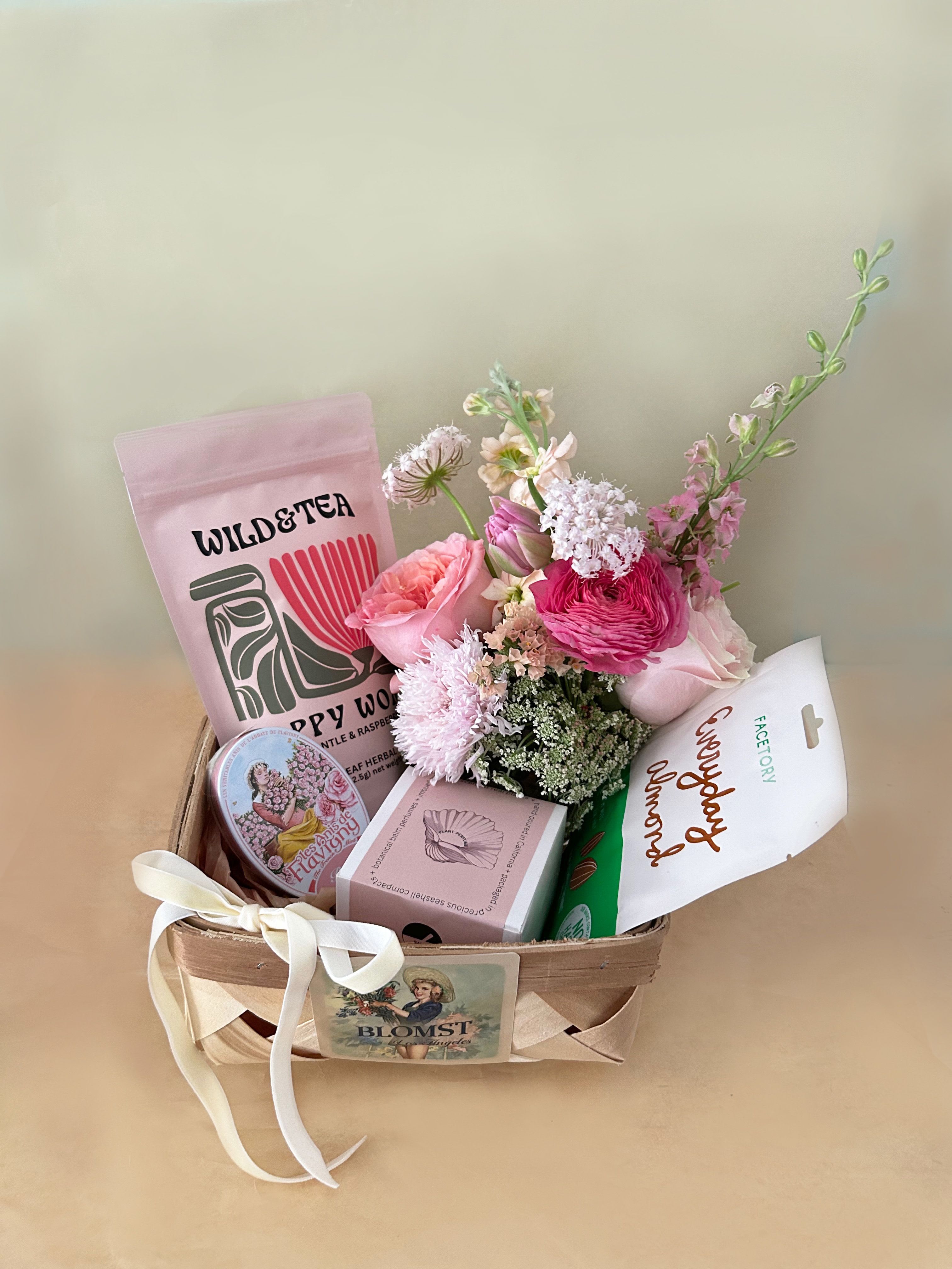 Vera Wellness Basket - A wellness basket in the pretty colors of pink, white and green in the flavors of jasmin, rose and herbs. The petite and classic wooden French basket measures 7" by 7" by 4" and is overflowing with lovely treats and flowers. The basket contains: Wild &amp; Tea herbal tea "Happy Womb": Enjoy a beautiful, herbal tea that reduces pms symptoms and strengthens your womb. Use during the last month of pregnancy to prepare for labor. Organic ingredients- Lady's mantle, raspberry leaf and lemon balm Earth Tu Face Jasmine Solid Perfume Balm in a real shell. A gorgeous blend of Jasmine Grandiflorum and Jasmine Sambac oils. This luxurious and deeply fragrant fusion offers an intoxicating aroma, while delivering the natural healing and therapeutic benefits inherent in its rich scent.  Everyday Almond sheet Face mask. Sweet Almond oil extract for dark spots and uneven complexion. Rehydrates as well. Les Anis De Flavigny: with a subtly minty taste. These all-natural candies are housed in a vintage decorative tin, with about 30 mints per tin. Each anise seed is coated in naturally-flavored sugar to produce round and smooth rose flavored mints that will delight the palate and senses. A mini Blomst arrangement of fresh, pretty stems in a clear cylinder vase 4" by 4". Velvet ribbon. ready for hand delivery in LA.