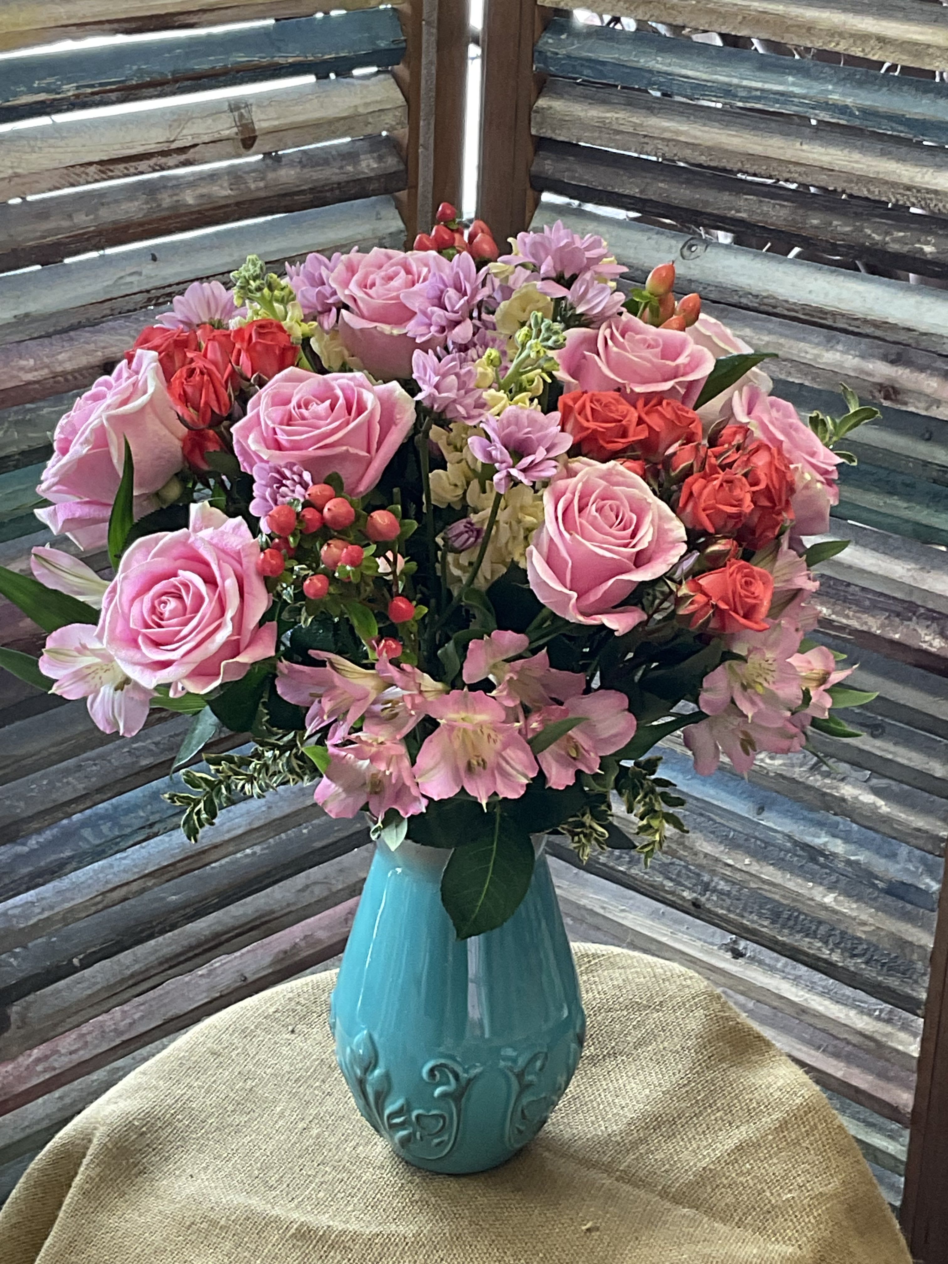 26-112 - A lush, rounded bouquet of pink roses, orange spray roses, and yellow stock paired with pale pink alstroemeria and soft lavender daisy-like blooms-this flower arrangement is an everyday gift arrangement that reads romantic and joyful at once. Perfect as a birthday bouquet, anniversary arrangement, Mother's Day gift, or a "just because" surprise, it's designed to make the person you're thinking of feel celebrated and seen with its warm, layered colors.  The mix includes roses, alstroemeria, daisies, and complementary filler blooms in shades of pink, red, purple, and white, tightly gathered for a full, abundant look. Flowers arrive arranged in a decorative turquoise ceramic vase that adds a bright, cheerful contrast and makes the piece ready to display. The compact, rounded shape keeps the composition neat on a table, mantel, or bedside.  This arrangement suits recipients who appreciate soft, romantic color palettes and a polished presentation-mothers, partners, close friends, or anyone who could use a thoughtful lift. Vase includes water source at delivery; trim stems and refresh water every few days to keep blooms looking their best.
