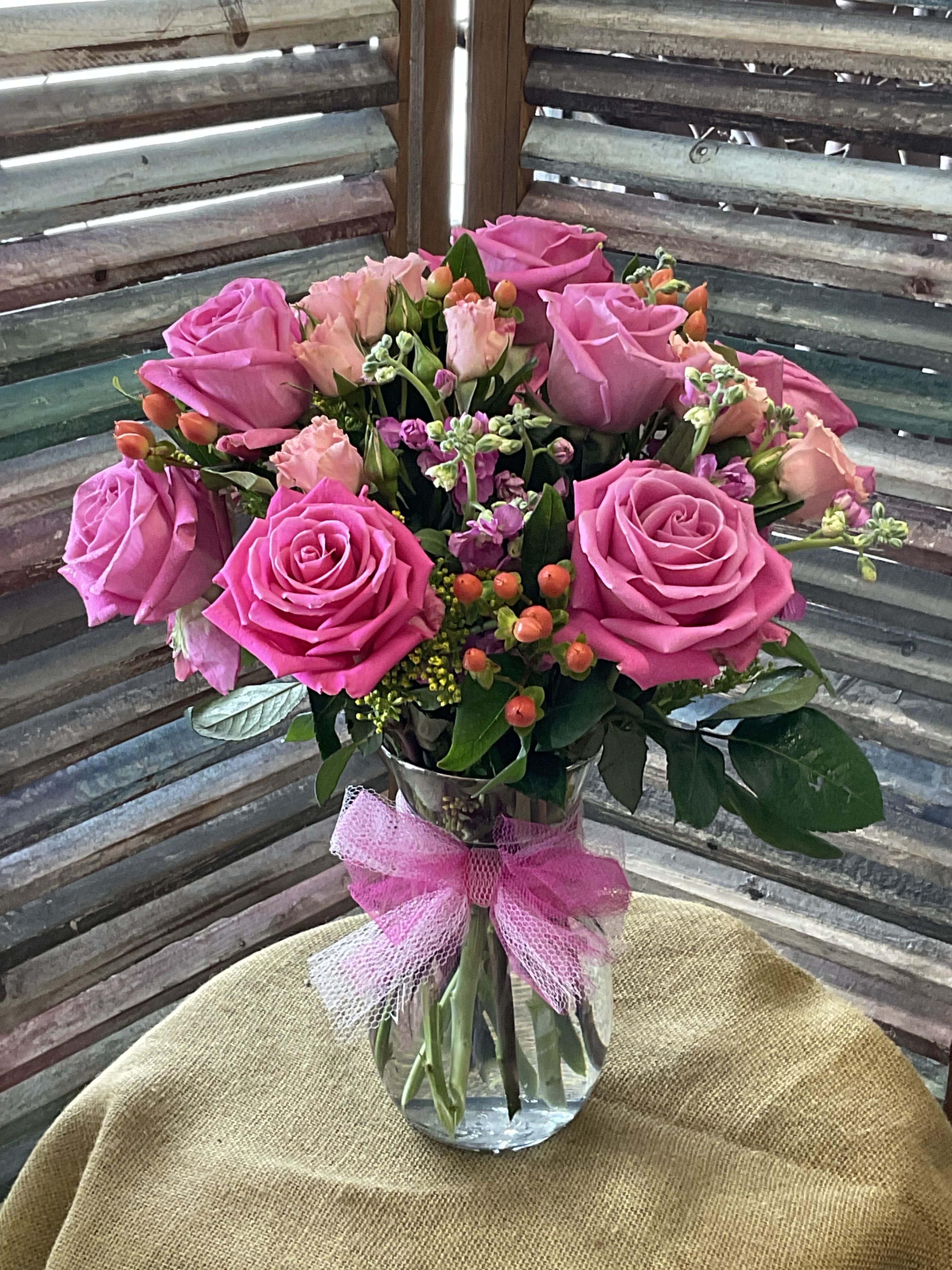 26-113 - Bright pink roses take center stage in this generous floral arrangement, perfect as a birthday bouquet, anniversary surprise, or a thoughtful thinking-of-you gift. The rounded, lush design reads as cheerful and romantic-she'll feel genuinely celebrated the moment these pink, green, and white blooms arrive.  A cluster of bright pink roses is paired with smaller pale-pink blossoms and tiny white accents for texture, all set in a clear glass vase and finished with a soft pink tulle ribbon at the neck. The compact, rounded shape makes it easy to place on a bedside table or dining room console; colors are purposely feminine and uplifting without being overly formal. For best enjoyment, keep fresh water in the vase and trim stems if you transfer them-this arrangement is designed to bring a fresh, joyful look to everyday gifting moments.