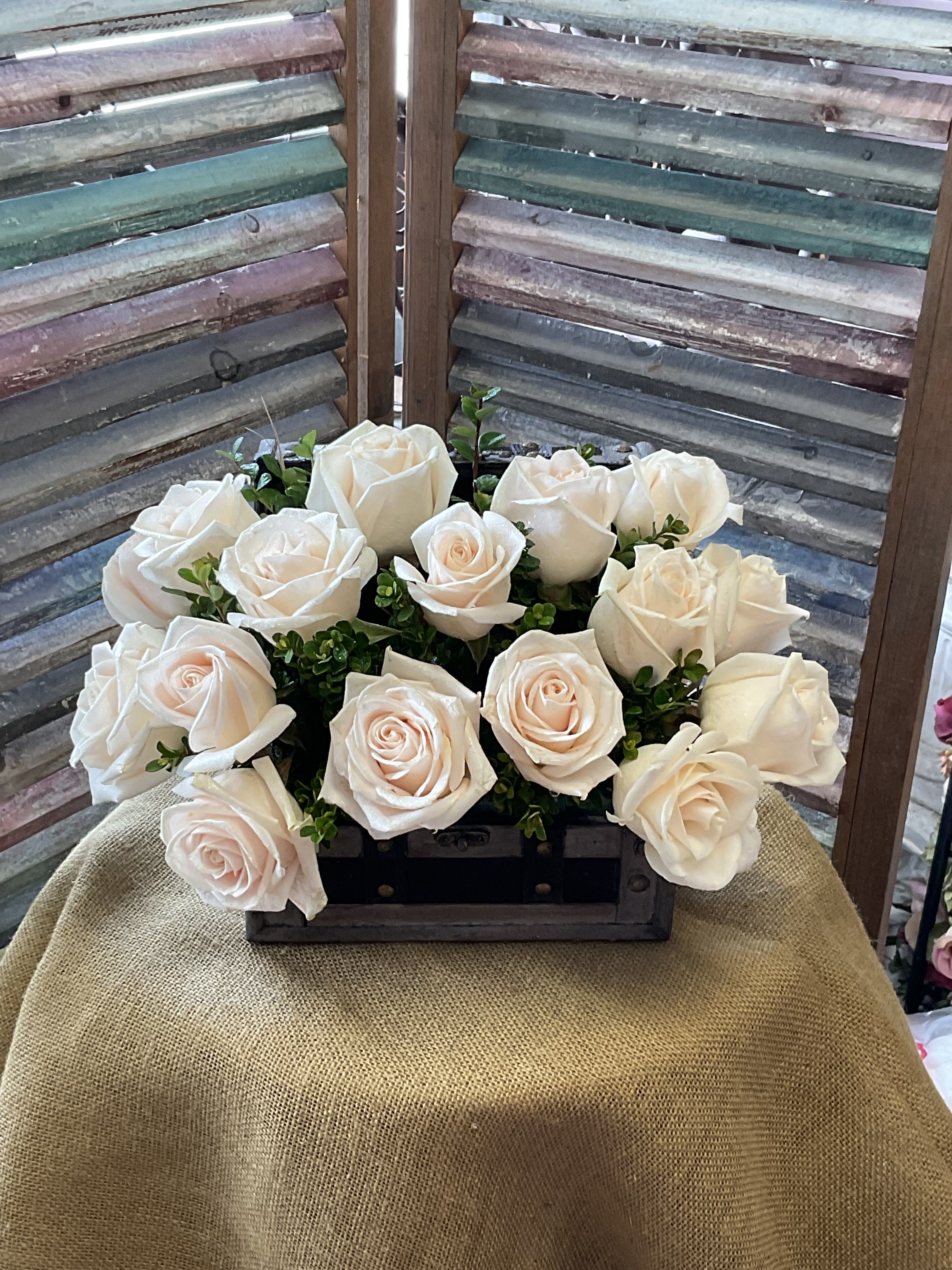 26-114 - A compact cluster of cream-to-white roses with a faint blush center creates a quietly elegant gift perfect for anniversary roses, sympathy arrangements, or a thoughtful everyday bouquet. This low, garden-like flower arrangement in a dark wooden box reads as calm and handcrafted-the kind of floral gift that makes the person you're thinking of feel soothed and truly seen.  Cream and soft-pink roses are packed closely together with small sprigs of green tucked between the blooms for delicate contrast and texture. The roses sit in a rustic wooden box that keeps the design low and full, making it ideal for tabletops or as a centerpiece without blocking conversation. The neutral palette and compact shape suit weddings, quiet celebrations, or moments when you want to offer comfort; care is simple-keep water fresh and display away from direct heat for longest vase life.