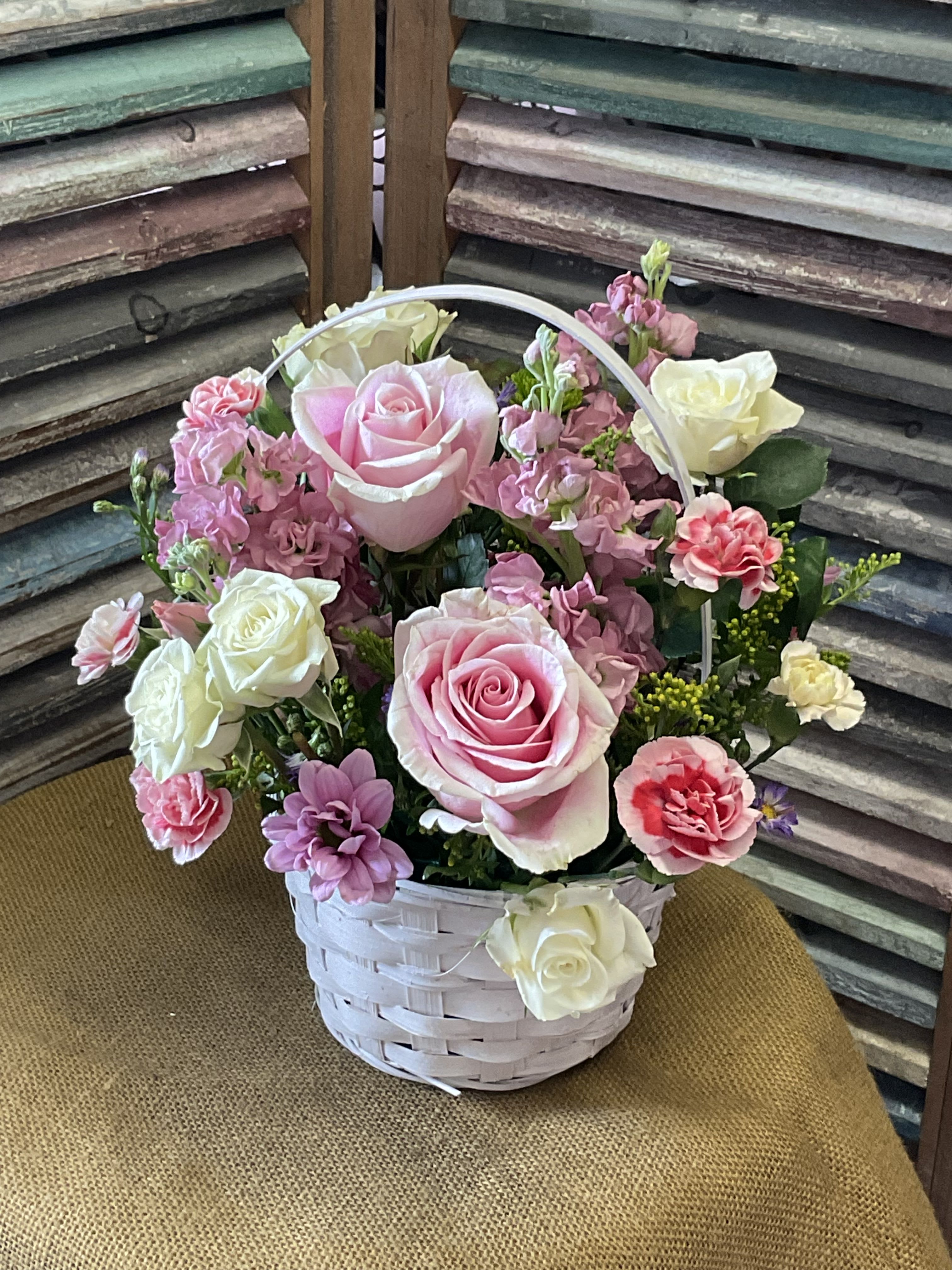 26-111 - A soft, garden-style arrangement of pink roses and white spray roses arranged with pink mini carnations and purple-hued stock creates an immediate impression of gentle romance-ideal as a birthday bouquet, anniversary flowers, Mother's Day gift, or a thank-you gesture. The white wicker basket with a curved handle gives the composition a charming, cottagelike feel that makes the recipient feel truly seen and celebrated the moment it arrives.  The arrangement is densely gathered for a full, rounded silhouette: lush roses anchor the design while small pink carnations and clustered stock provide delicate texture and subtle purple accents. Set in a white wicker basket, the piece reads cheerful and feminine and is ready to display-no additional vase needed. This is the kind of gift that conveys warmth and appreciation, perfect for the person you want to brighten with a touch of floral charm.