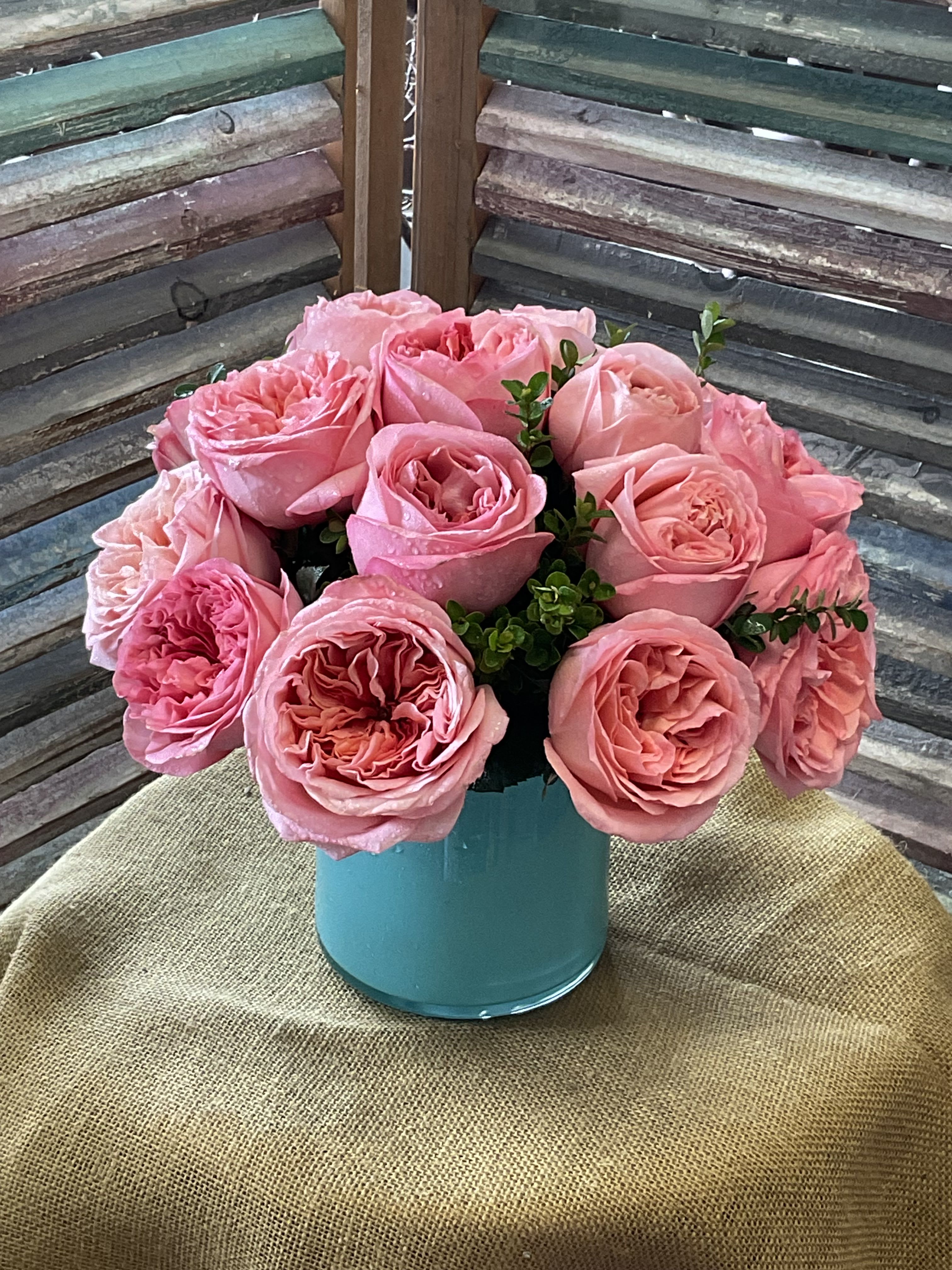 26-115 - A lush dome of pink garden roses makes an immediate, romantic impression in this flower arrangement-perfect as a birthday bouquet, anniversary gift, or a heartfelt Mother's Day surprise. The rounded cluster of full, layered pink rose petals reads soft and feminine, giving the person you're thinking of a fresh garden-picked moment that feels both intimate and celebratory.  The roses are arranged tightly in a smooth turquoise ceramic vase that brightens the blooms and adds a cheerful pop of blue-green. Small sprigs of greenery peek through the bouquet. Sized to sit nicely on a tabletop or bedside, it's an easy way to make someone feel seen and loved-no fuss care keeps the roses looking their best for days.