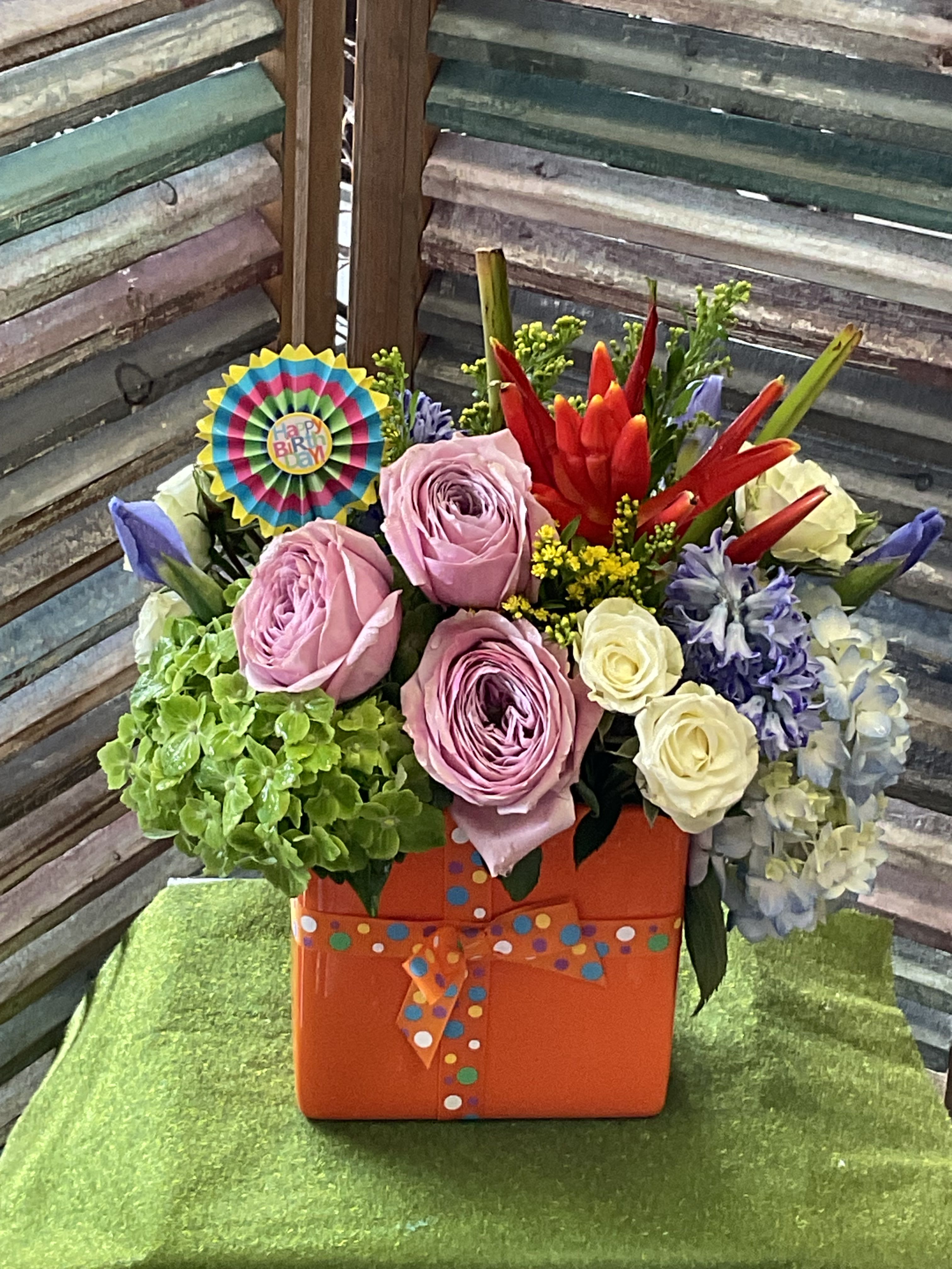 26-116 - Bold, cheerful and undeniably festive, this birthday flower arrangement is made to make someone feel celebrated the moment they see it. A bright mix of blush-pink garden roses, and white spray roses paired with a vivid green hydrangea cluster, pops of purple blooms and striking red tropical-style flowers create a lively, textured display that reads joyful and abundant-a perfect birthday bouquet or celebratory everyday gift for someone who loves color.  Arranged in a square orange gift box and finished with a multicolored polka-dot ribbon, the presentation feels playful and ready to gift; a round birthday pick tucked into the design makes the occasion clear. The combination of large roses and voluminous hydrangea gives the arrangement a full, luxurious shape, while the contrasting red and purple accents add depth and energy.  Choose this arrangement when you want the recipient to feel truly noticed-bright, fun, and festive for birthdays, congratulations, or just-because surprises. The sturdy gift box makes it easy to display right away; order with confidence for same-week celebrations and expect a bold, colorful statement that will brighten any room.
