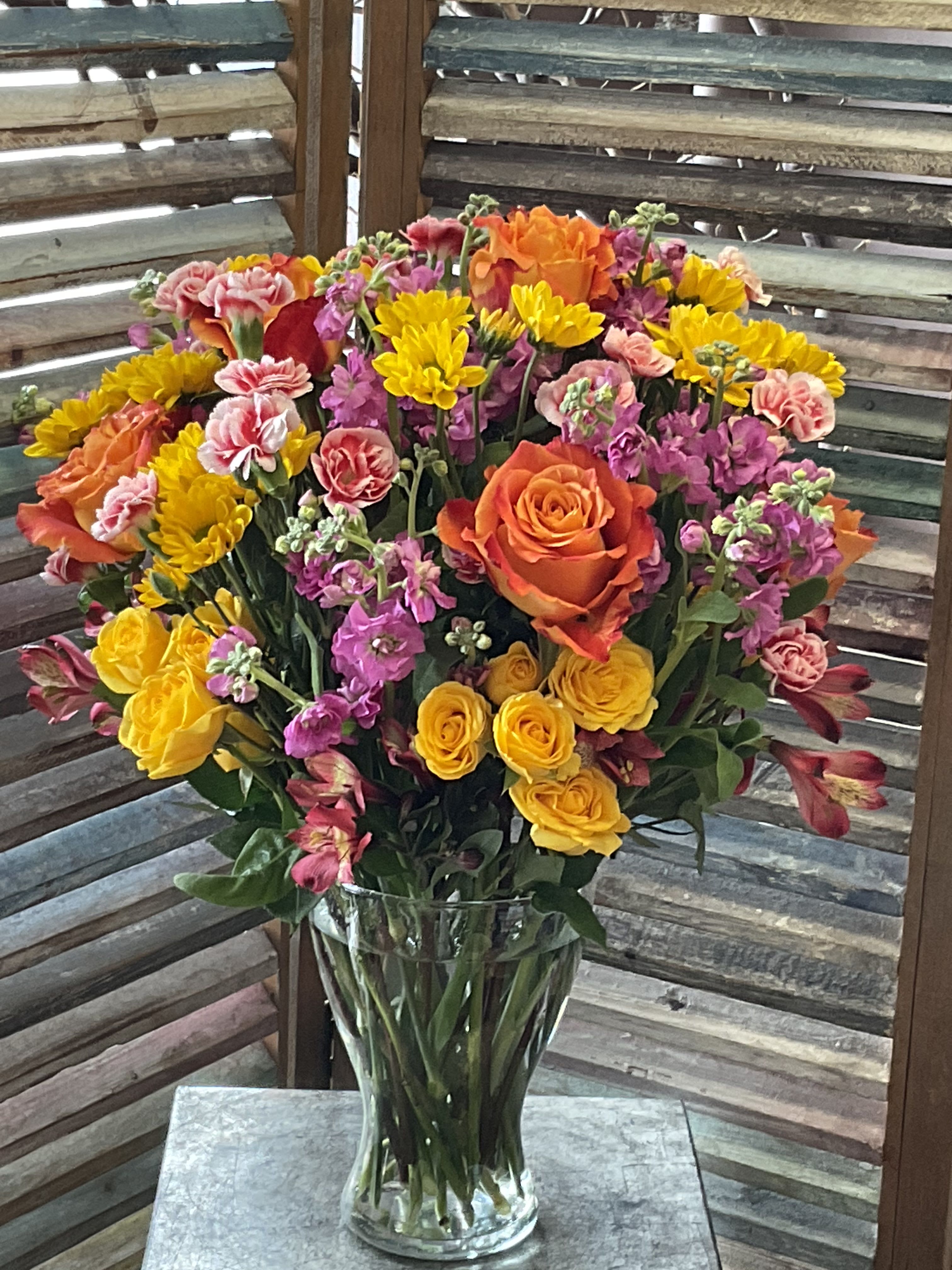 26-118 - Bright, cheerful and abundantly floral, this mixed-color flower arrangement is an everyday celebratory bouquet made to brighten birthdays, say thank you, or celebrate a springtime moment. The full, rounded design in a clear glass vase puts orange roses and yellow spray roses center stage, with pink carnations, purple stock, and pink alstroemeria woven through for playful contrast-an arrangement that reads happy, warm, and effortlessly festive for the person you're thinking of.  Dense clusters of blooms create layered texture and a generous silhouette that looks great on a table, desk, or entryway. The palette of saturated yellow and orange warmed by pinks and purple gives both high visual impact and soft romantic notes; stems arrive arranged in a clear glass vase for immediate display. Send this when you want someone to feel genuinely celebrated-it's the kind of bouquet that brings a smile and fills a room with color.