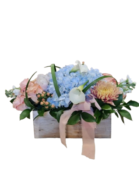 Seaside Garden Charm - Bring the beauty of a coastal garden indoors with the Seaside Garden Charm arrangement. Designed in a natural wooden box, this lush display features soft blue hydrangea, creamy white calla lilies, romantic blush roses, and a stunning focal bloom, all accented with fresh greenery and delicate textural elements.  Finished with a soft blush ribbon, this arrangement blends rustic charm with elegant coastal style—making it a perfect gift for birthdays, Mother’s Day, thank yous, or simply to brighten someone’s day.