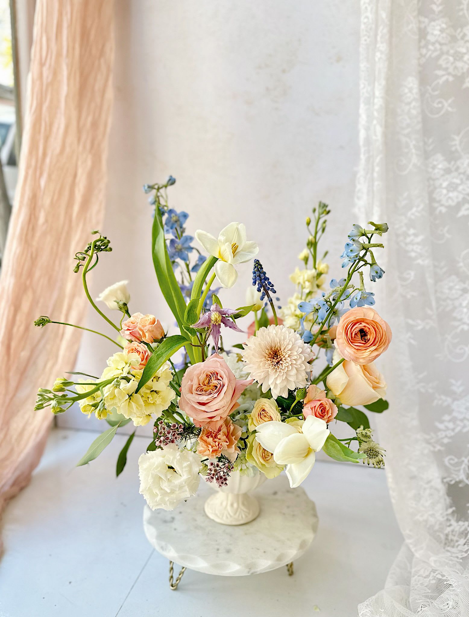 Golden Hour - A stunning vase arrangement featuring soft, peachy and white flowers, including roses, ranunculus, mums, tulips and touches of blue delphinium.  The arrangement will be designed in a ceramic compote vase.